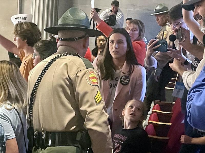 A woman standing next to a child looks a police officer in the eyes while talking and a large group of people are protesting in the background.