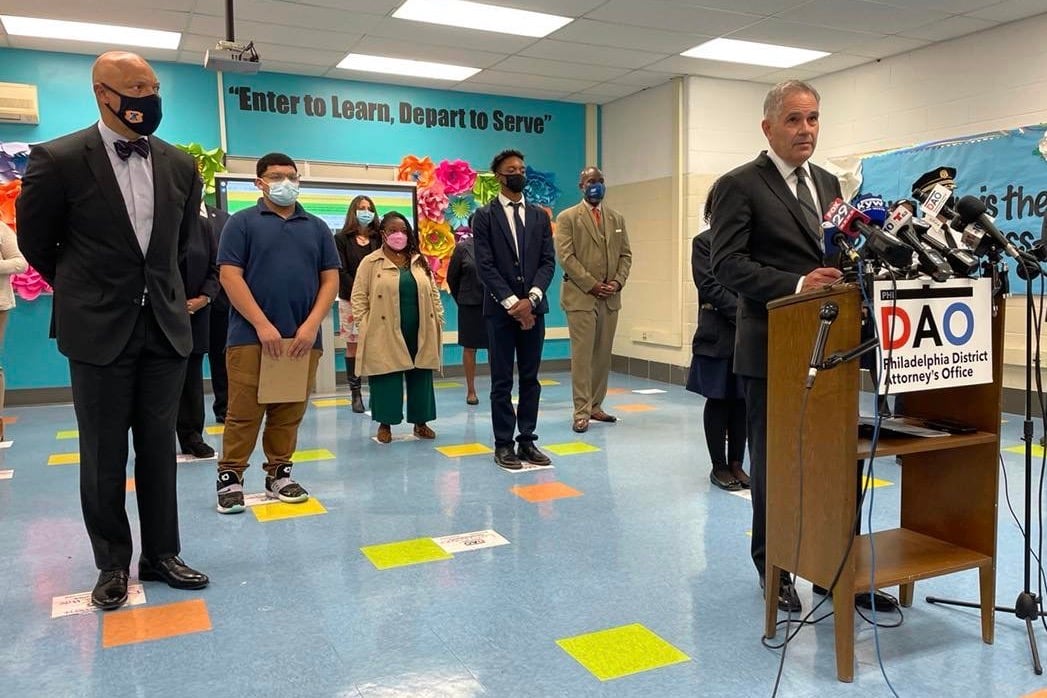 Philadelphia District Attorney Larry Krasner is speaking at a podium surrounded by microphones with people scattered, standing behind him.