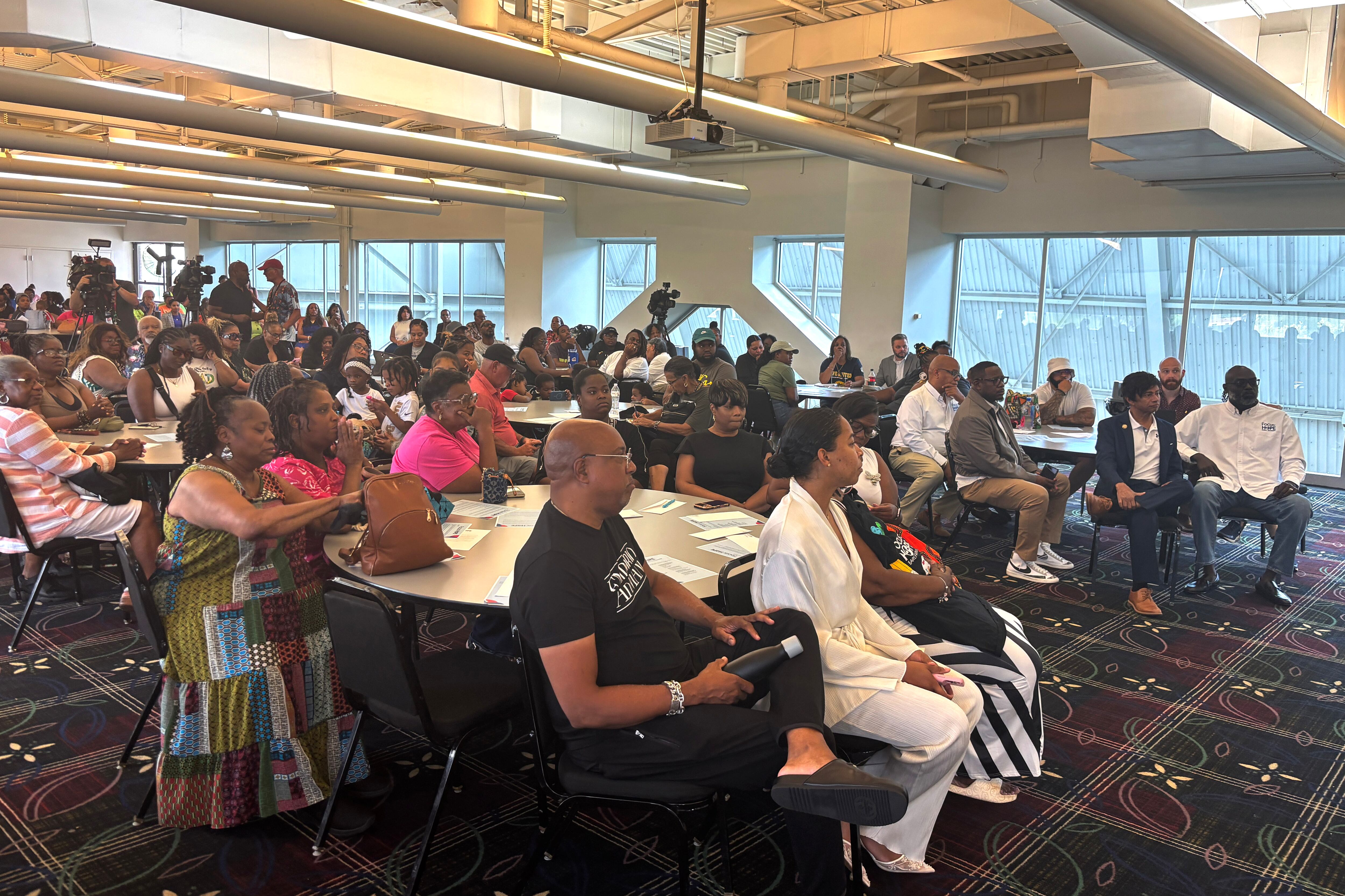 A crowd of people sits in a big room with a wall of windows.