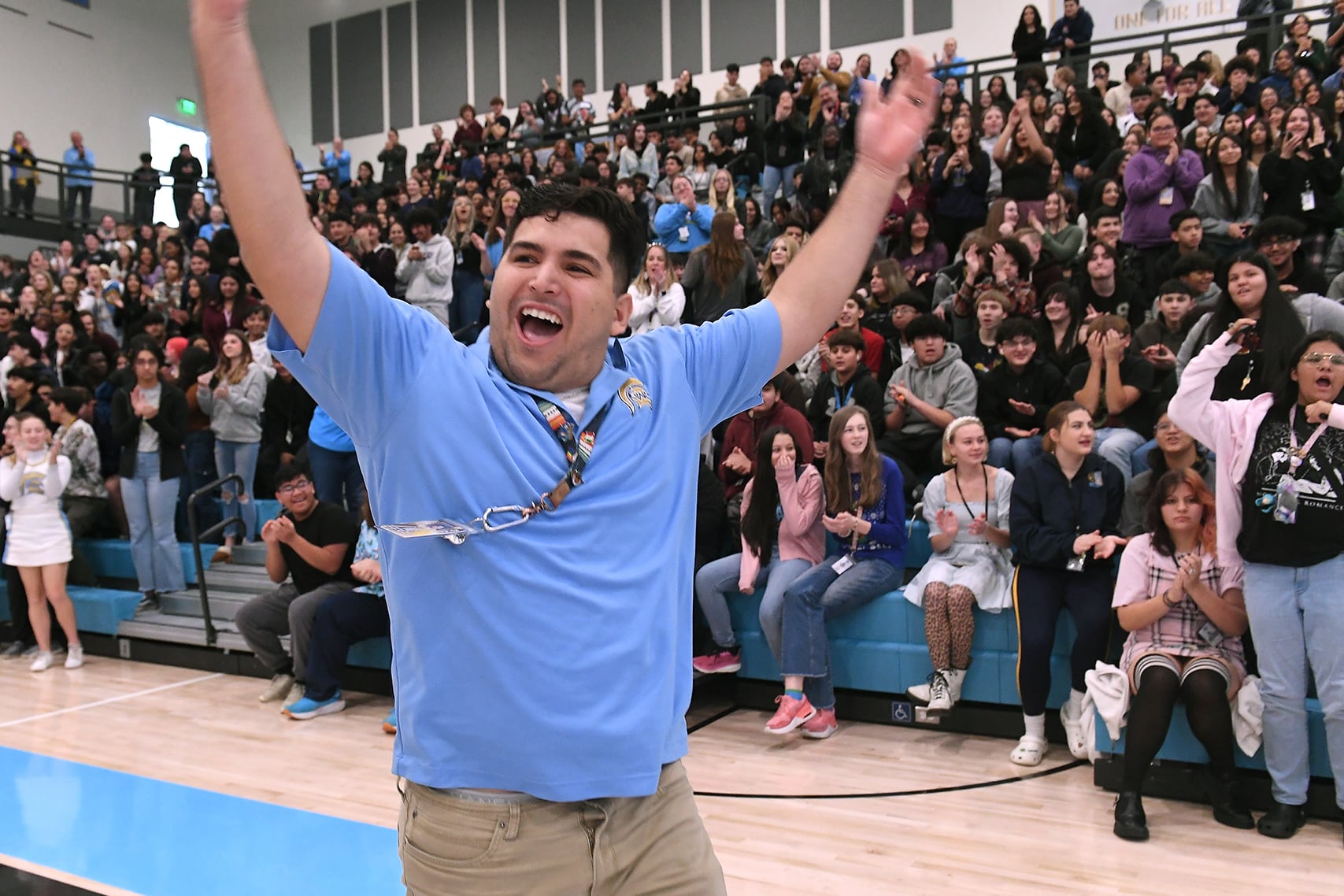 A man with short dark hair and wearing a blue t-shirt, waves his arms in the air while celebrating in a school gym with the bleachers full of people in the background.