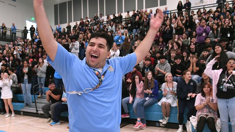 A man with short dark hair and wearing a blue t-shirt, waves his arms in the air while celebrating in a school gym with the bleachers full of people in the background.