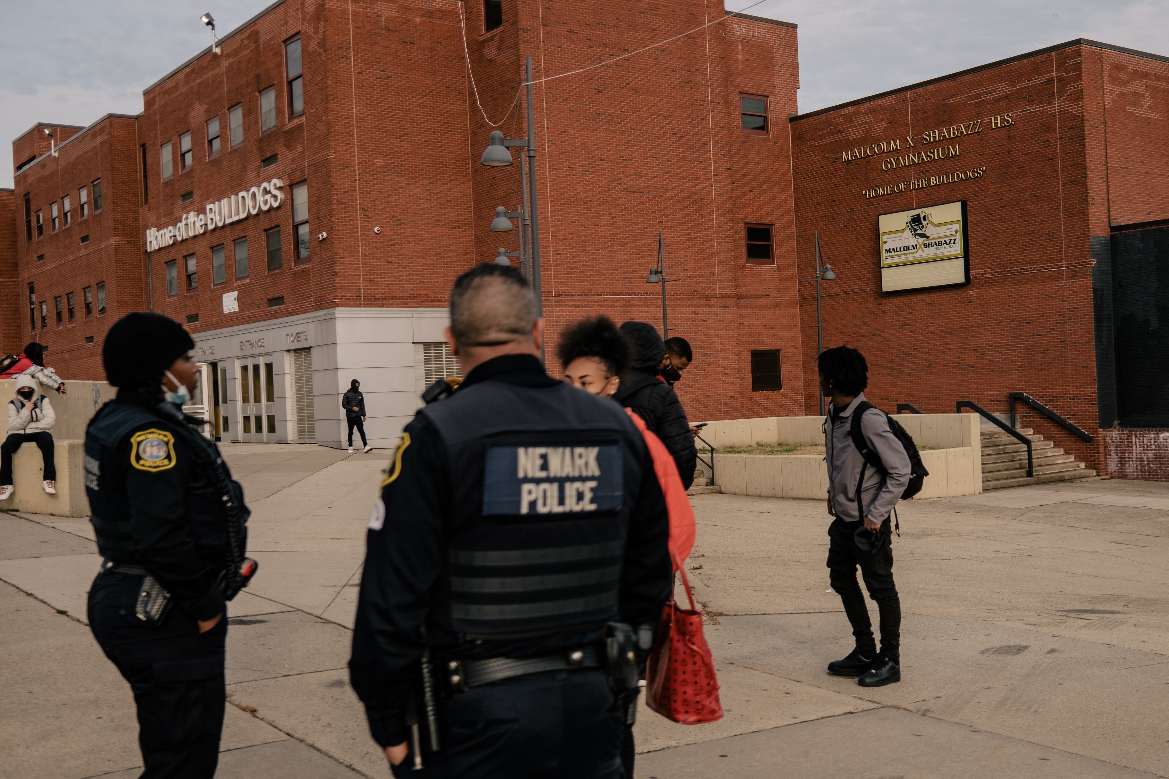 Members of the Newark Police department speak with a student outside of a large red-brick school front after dismissal, as other students wait outside of the school.