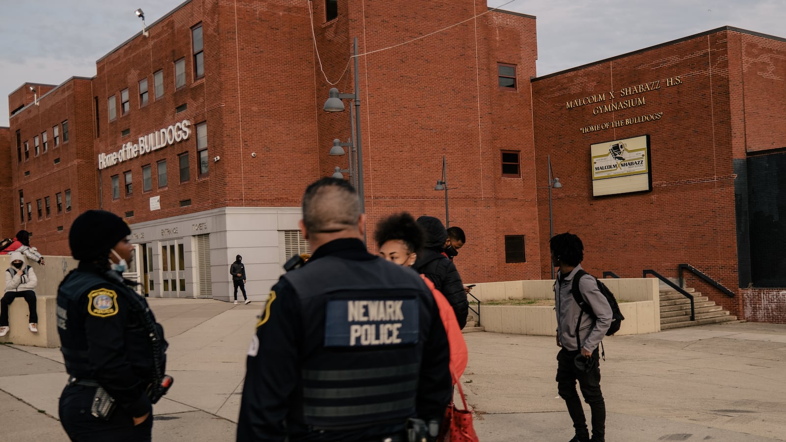 Members of the Newark Police department speak with a student outside of a large red-brick school front after dismissal, as other students wait outside of the school.