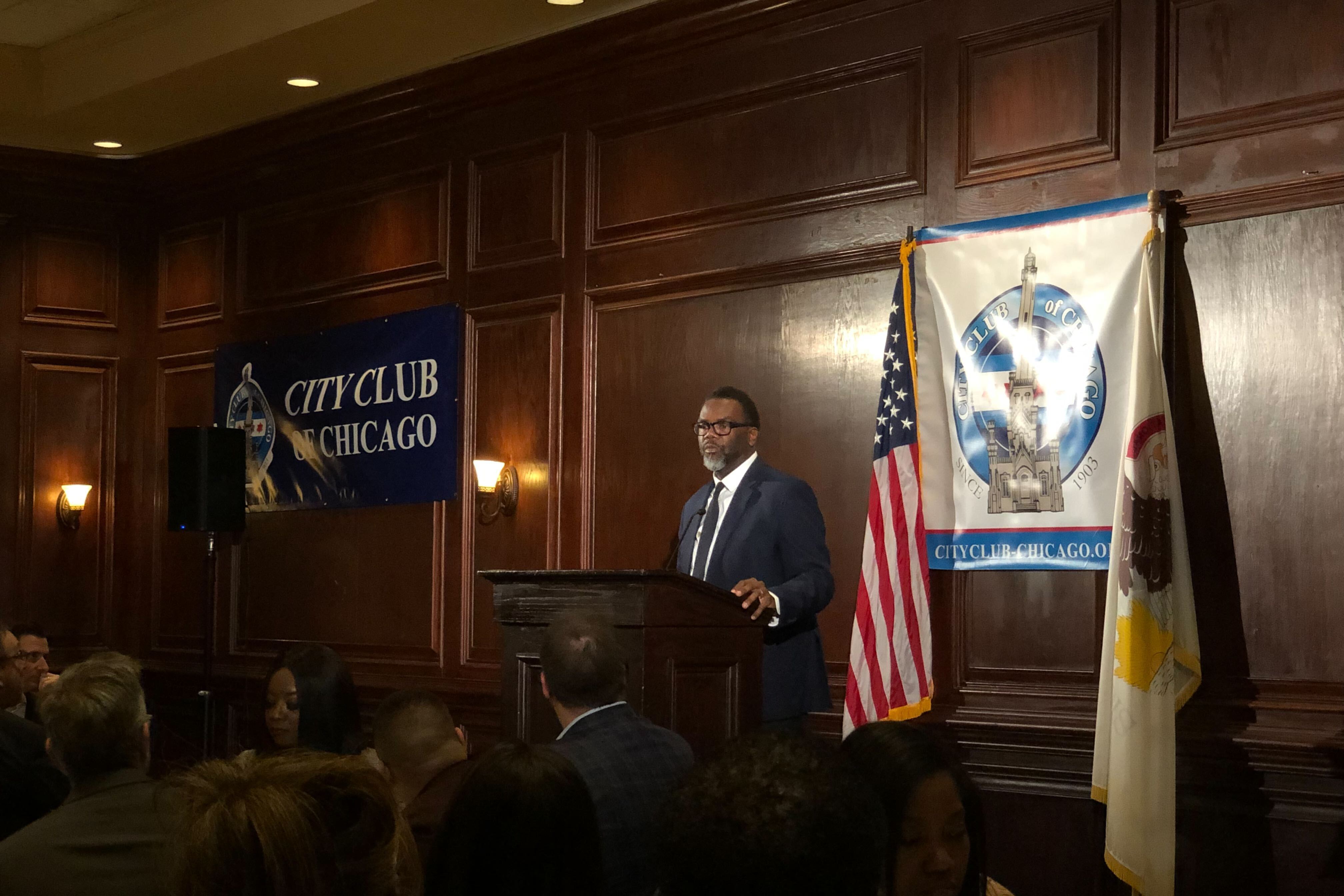 A stands at a lectern in front of a ballroom while a crowd of supporters watch from dining room tables.