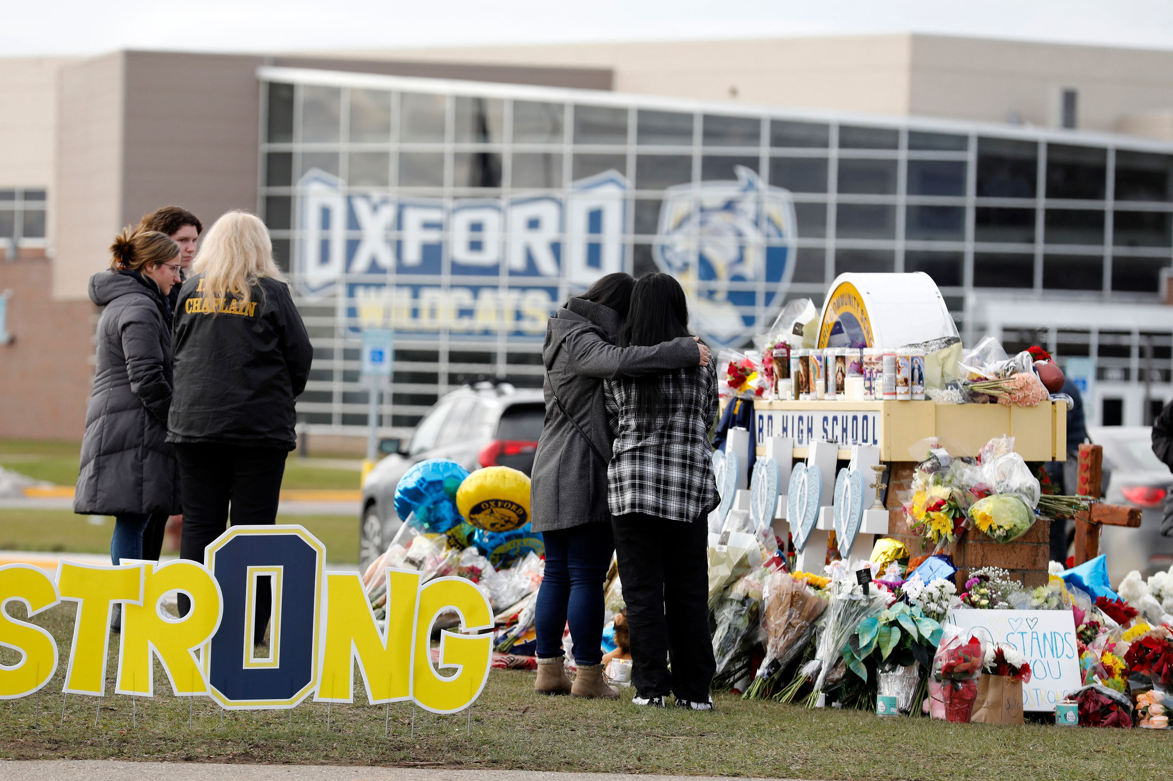 Two people wearing cold weather clothing embrace in front of a makeshift memorial in front of a large high school building while a group of three adults stand nearby.
