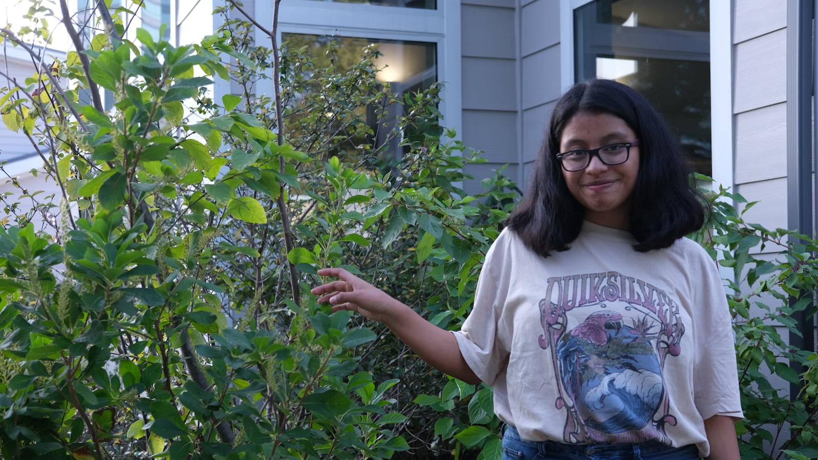 A teenage girl stands in front of plants/trees. She wears a beige shirt and blue jeans.