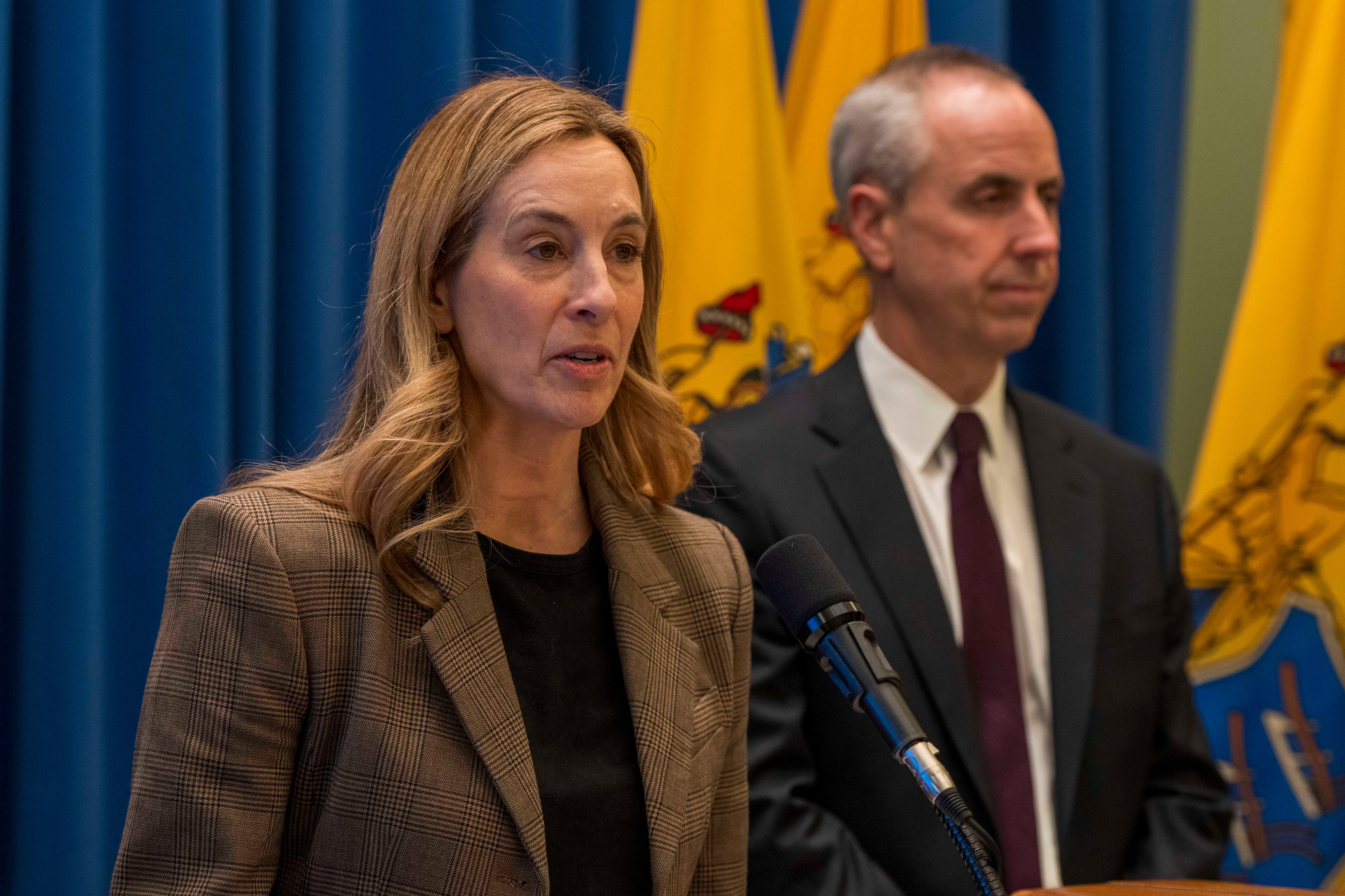 A woman in a brown jacked and black top speaks into a microphone.