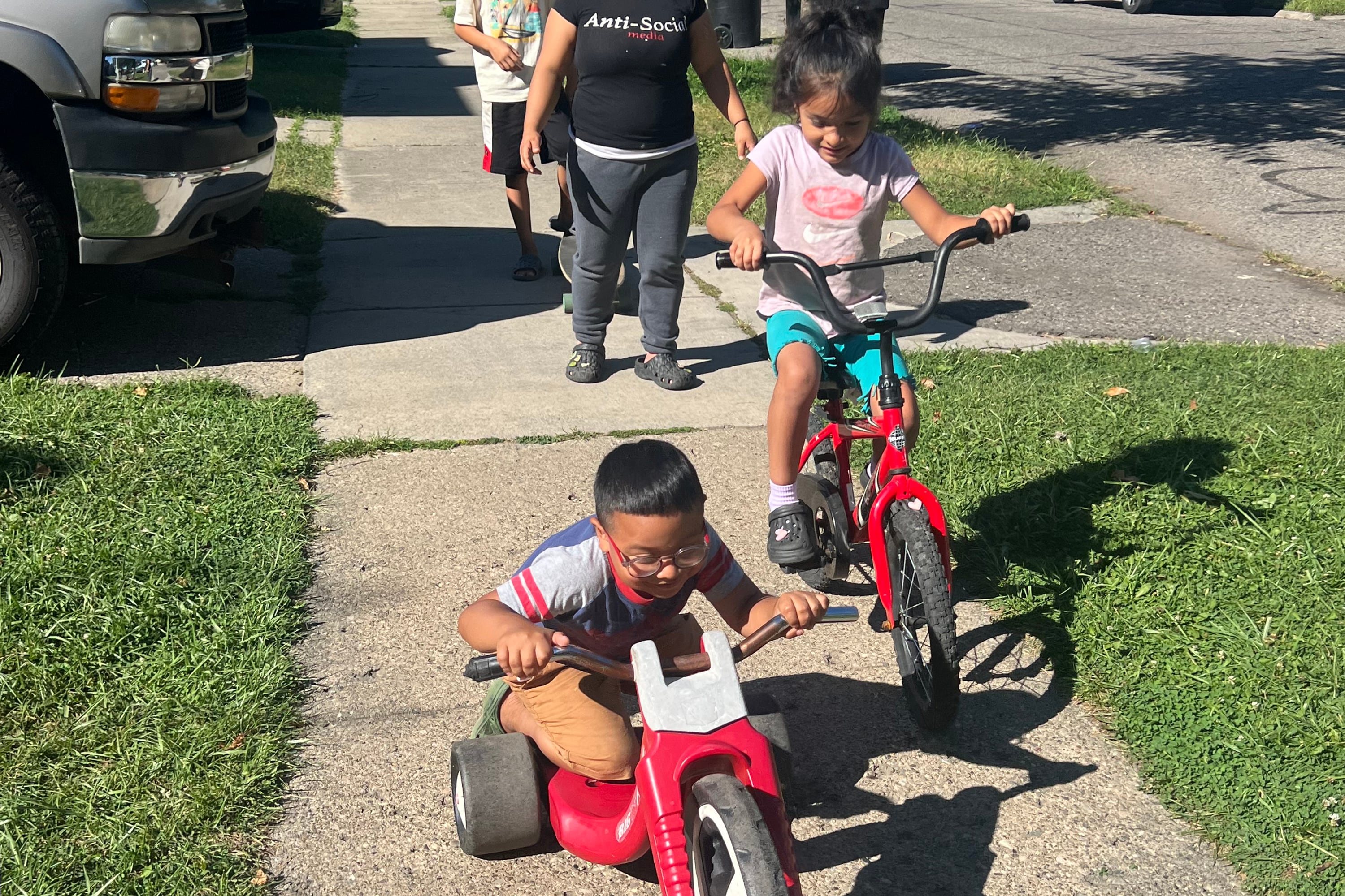 An adult and three kids walk and ride bikes down a sidewalk in a neighborhood.