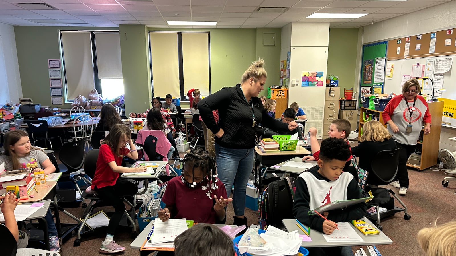 A woman in jeans and a black top with blonde hair stands in the middle of a classroom where several children are sitting at desks.