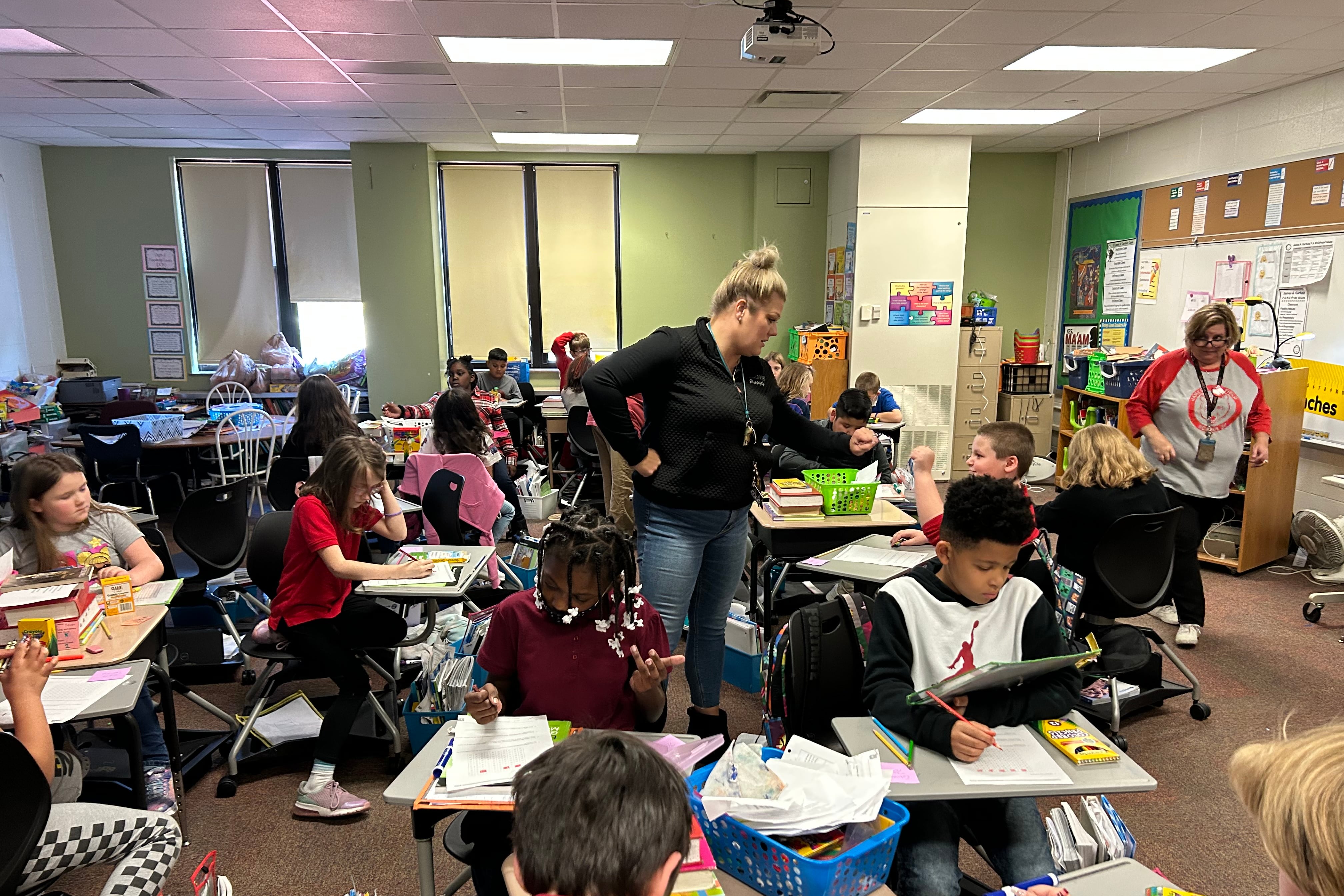 A woman in jeans and a black top with blonde hair stands in the middle of a classroom where several children are sitting at desks.