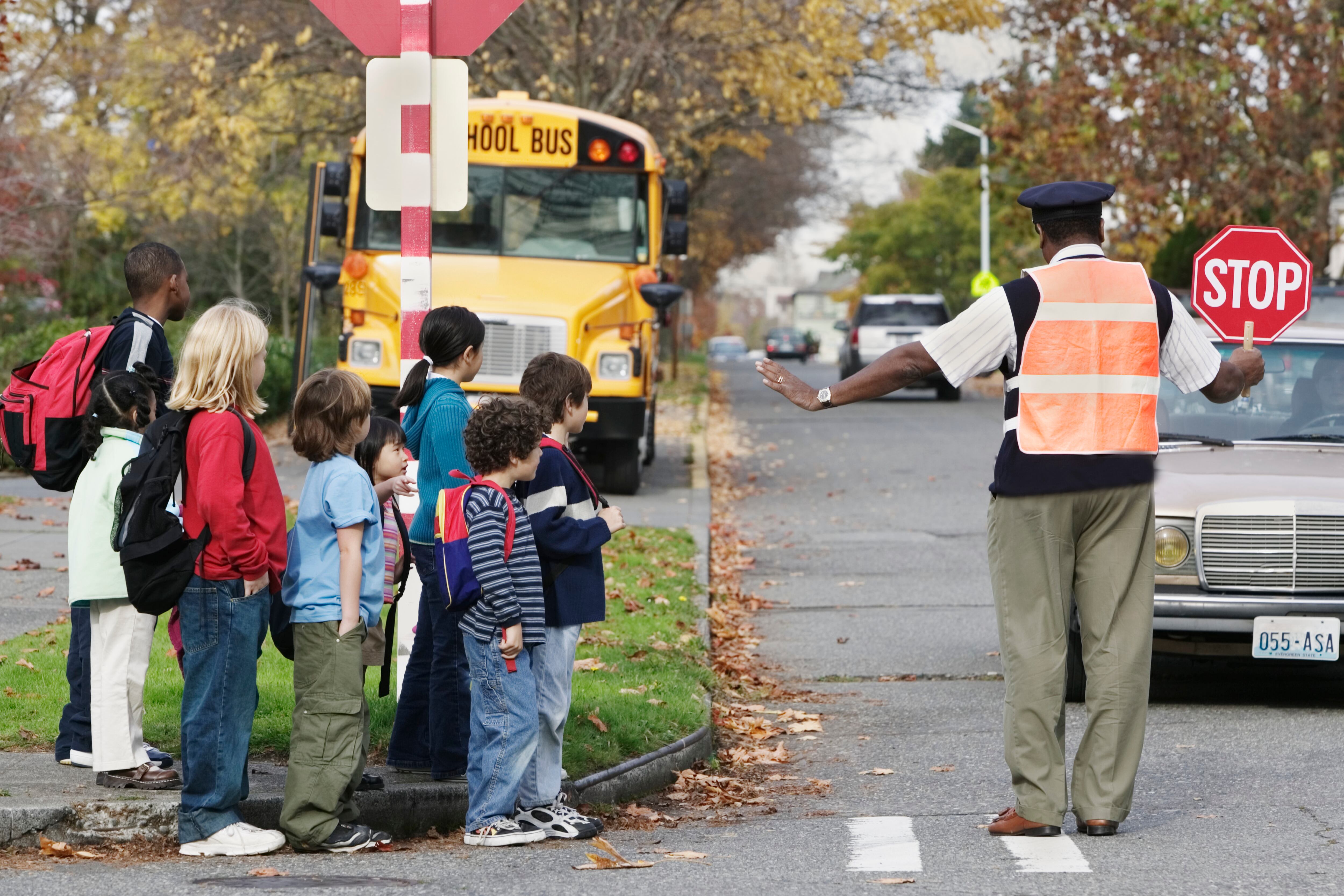 A man holds a stop sign and keeps students back from the street with a school bus in the background