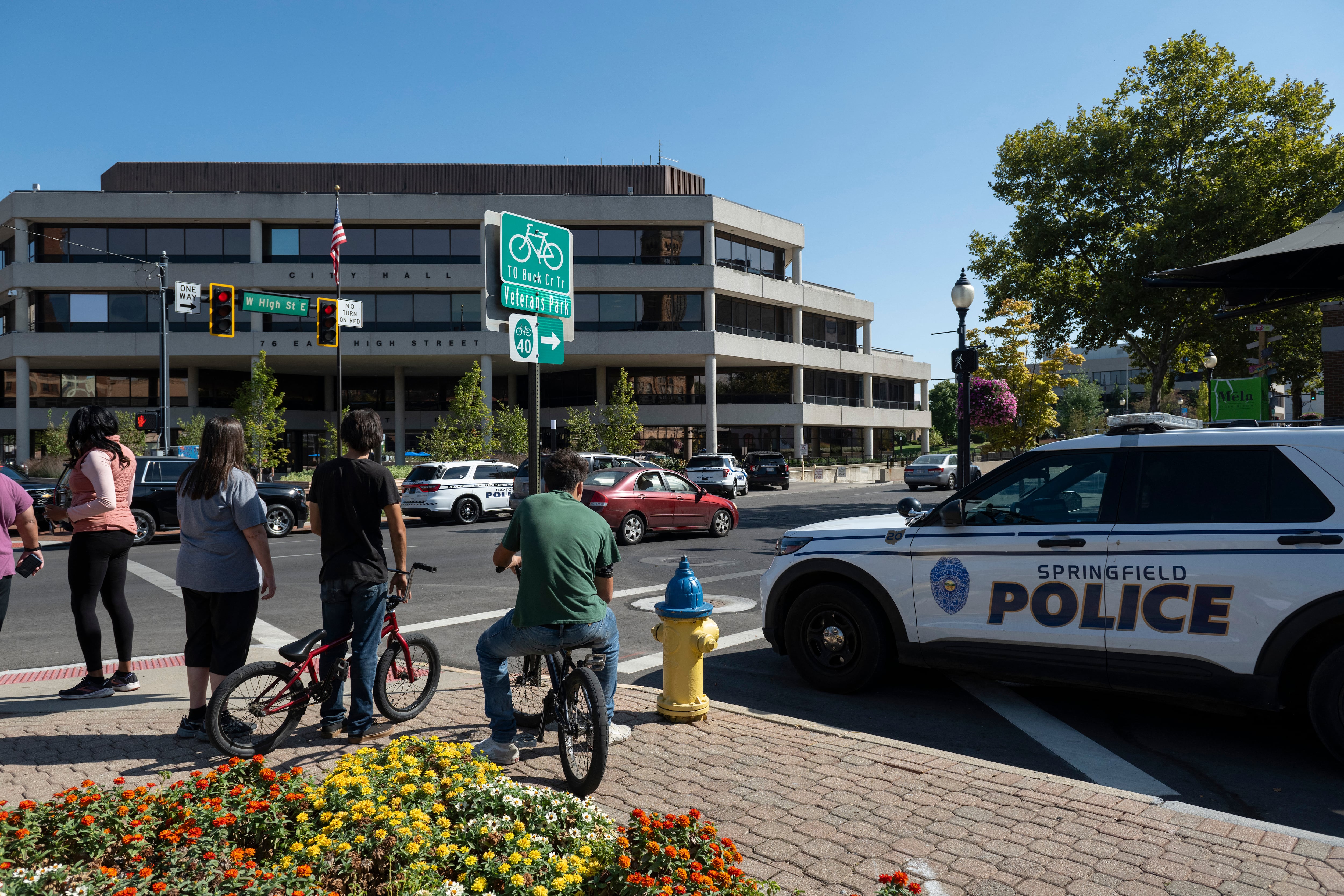 A group of people stand near a police car with a large building and blue sky in the background.