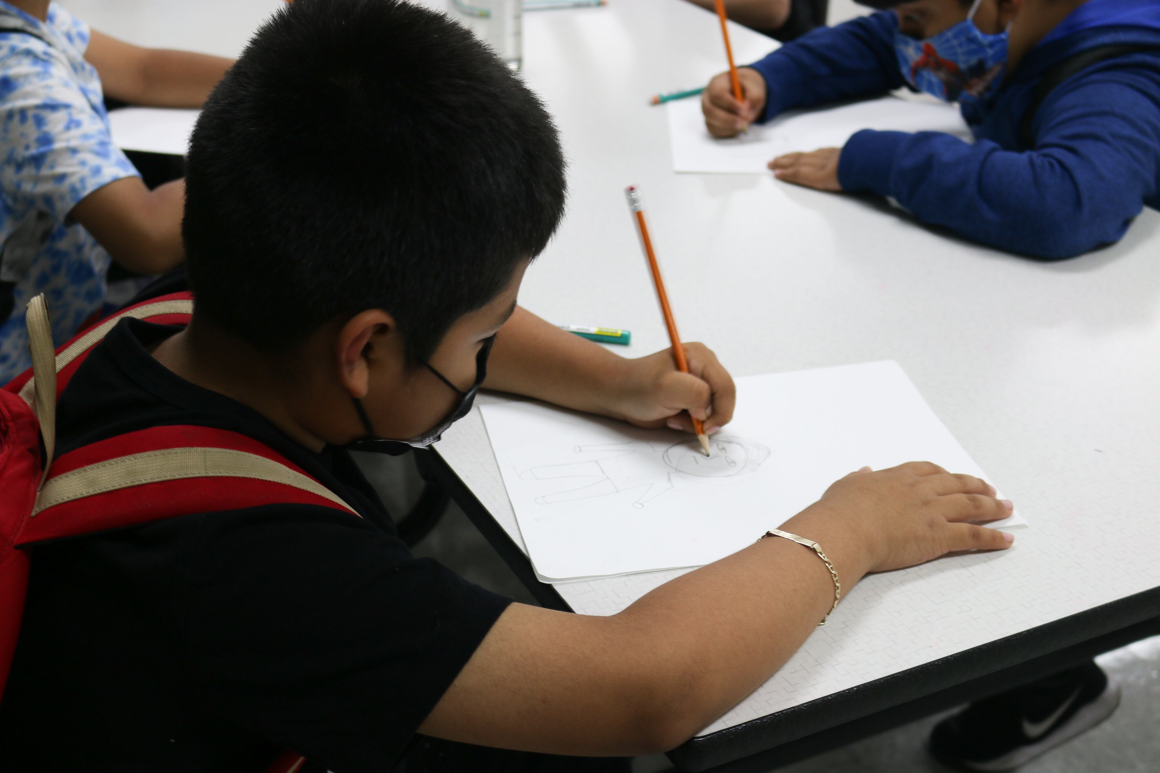 A child with short dark hair and wearing a mask writes with a pencil while seated as a desk.