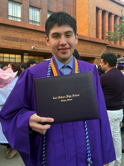 A photograph of a high school student posing for a photograph with his diploma and wearing a graduation gown outside of a high school building.