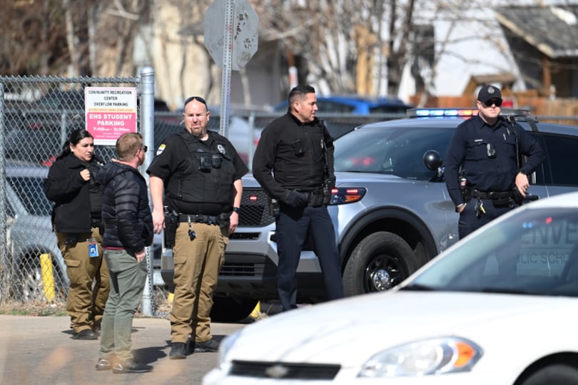 At least three police officers in uniform stand in a parking lot talking to two others. A white car is in the foreground.