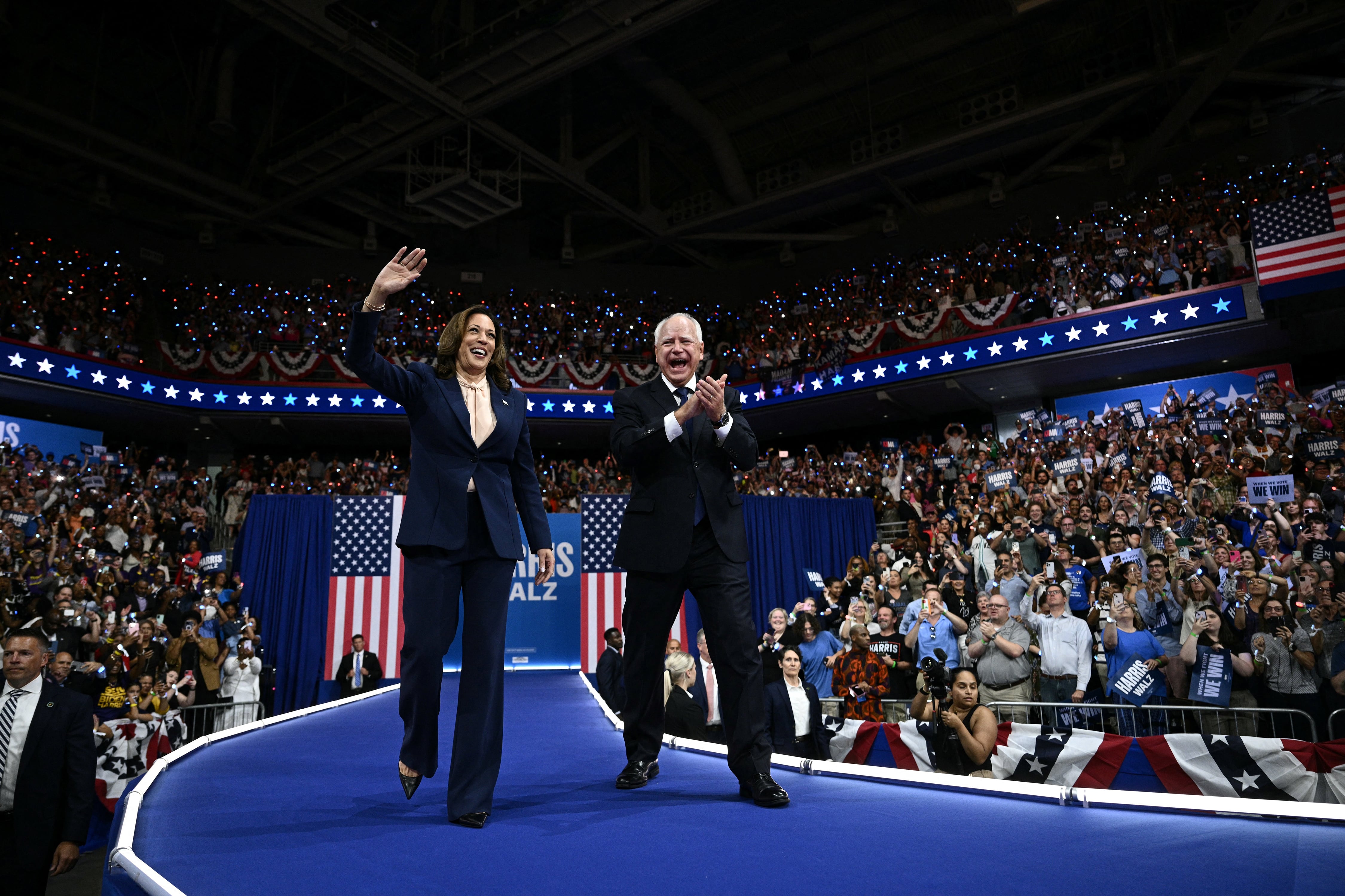A woman and man both wearing dark suits stand next to each other waving and clapping down a blue carpet walkway with a large crowd of people in the background.