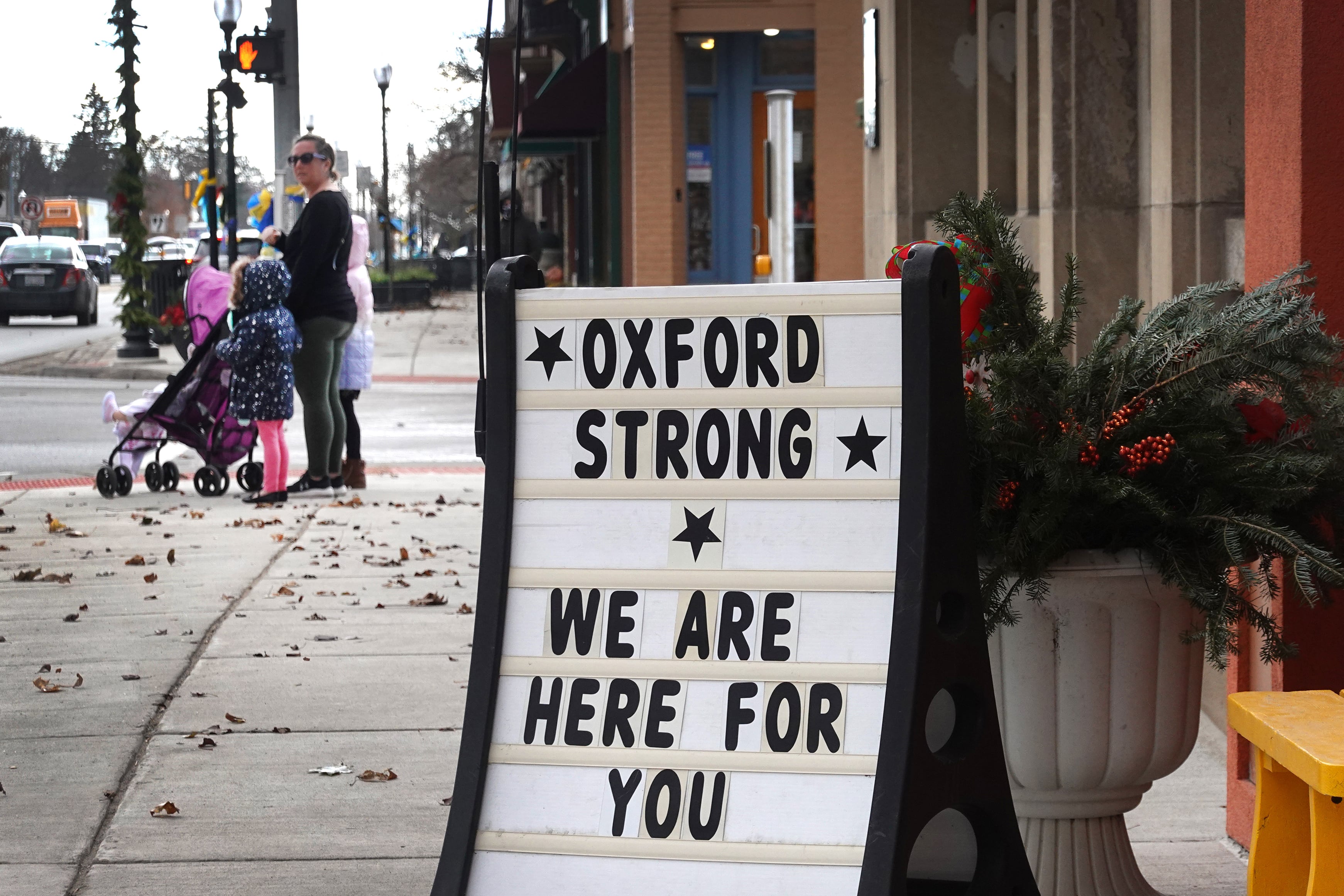 A white sign with black letters says "Oxford Strong We Are Here For You"  on a sidewalk with a family walking by in the background.