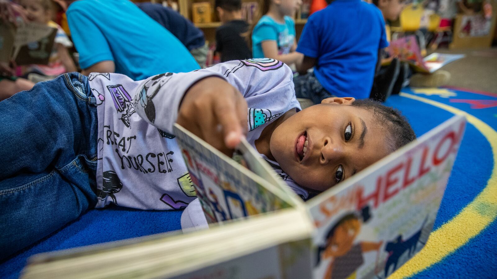 Five-year-old reads a book alongside lying on a rug alongside her classmates.