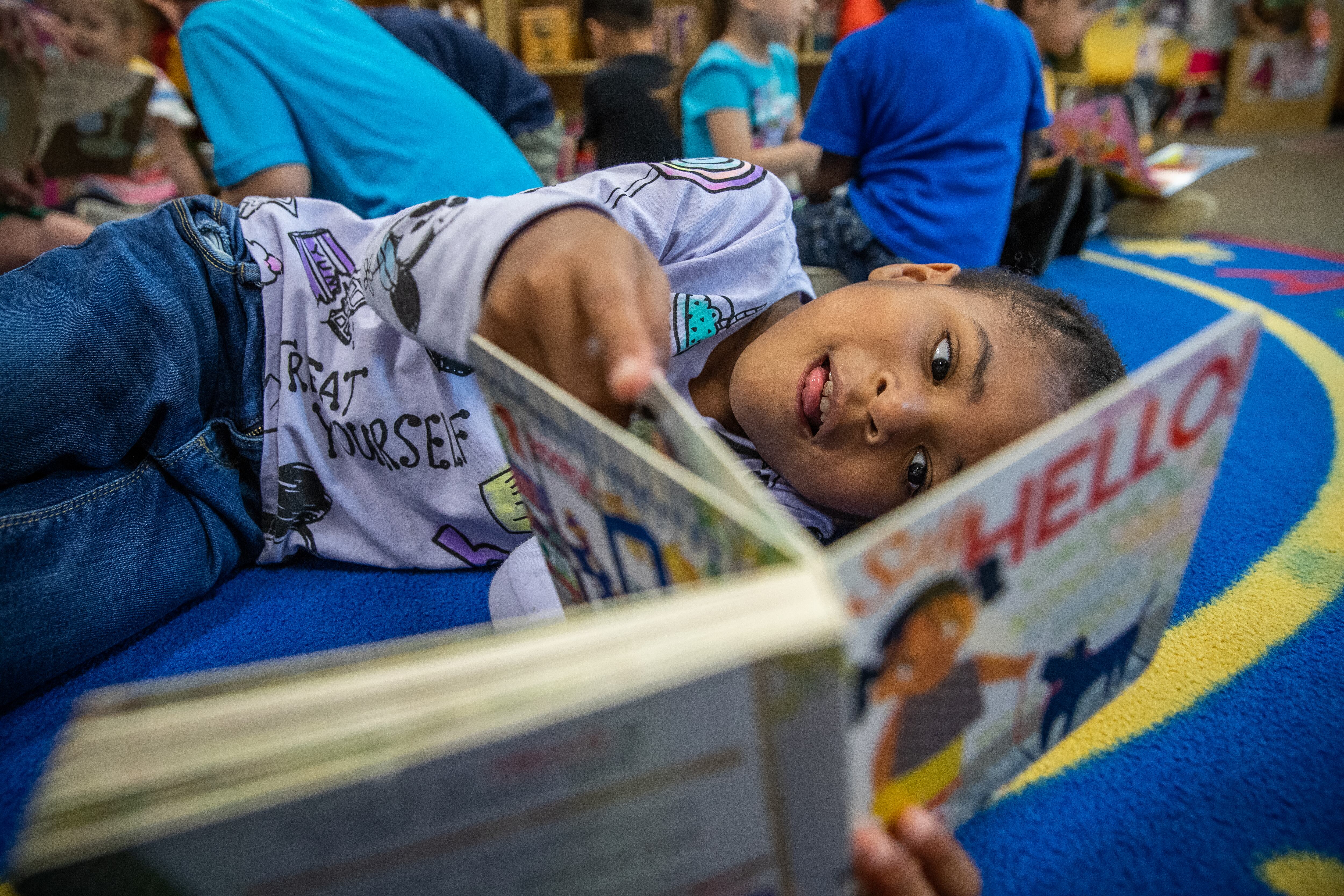Five-year-old reads a book alongside lying on a rug alongside her classmates.