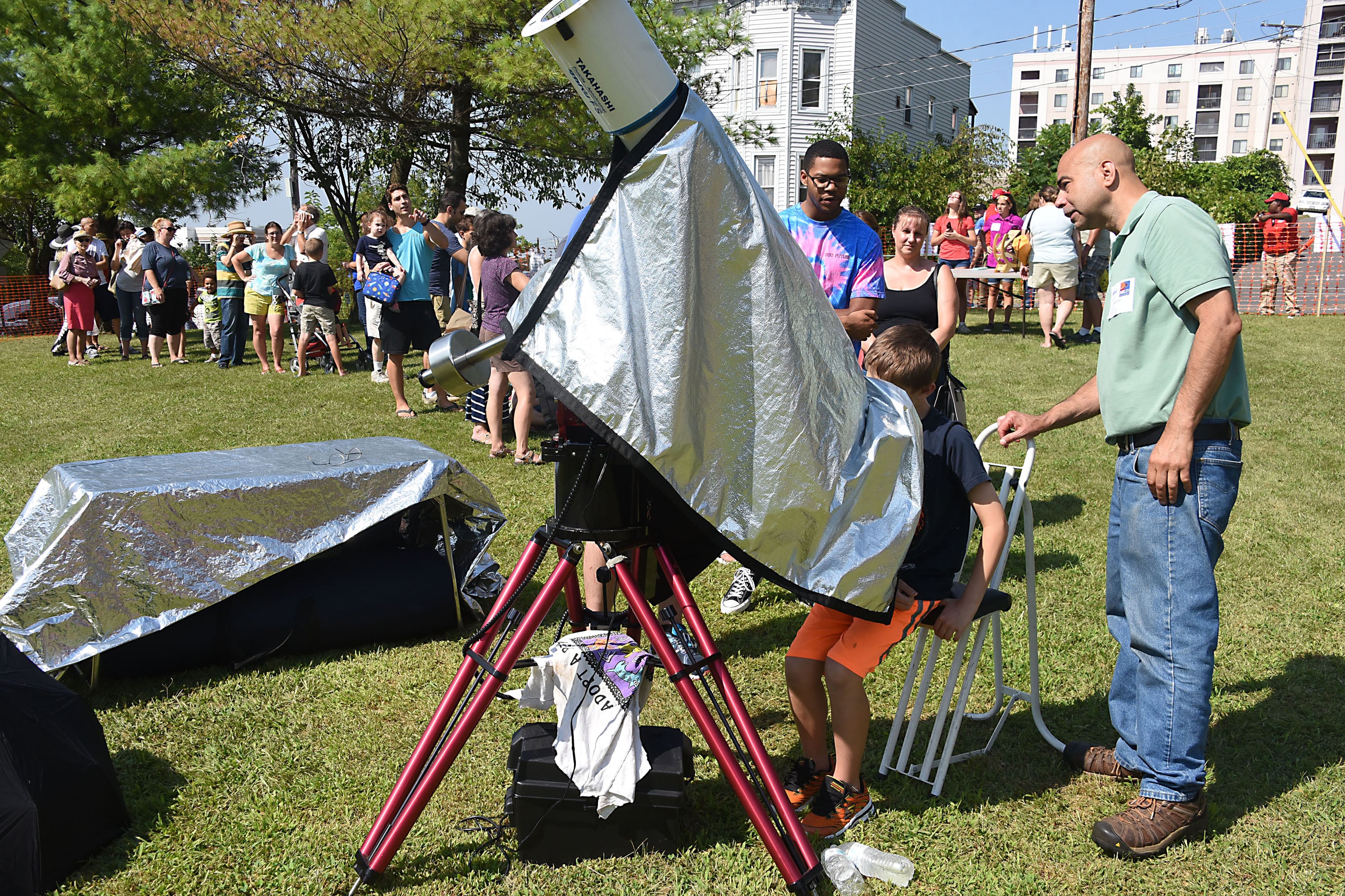 A long line of young people stand outside with one young person and an adult look through a telescope at the sun before an eclipse.