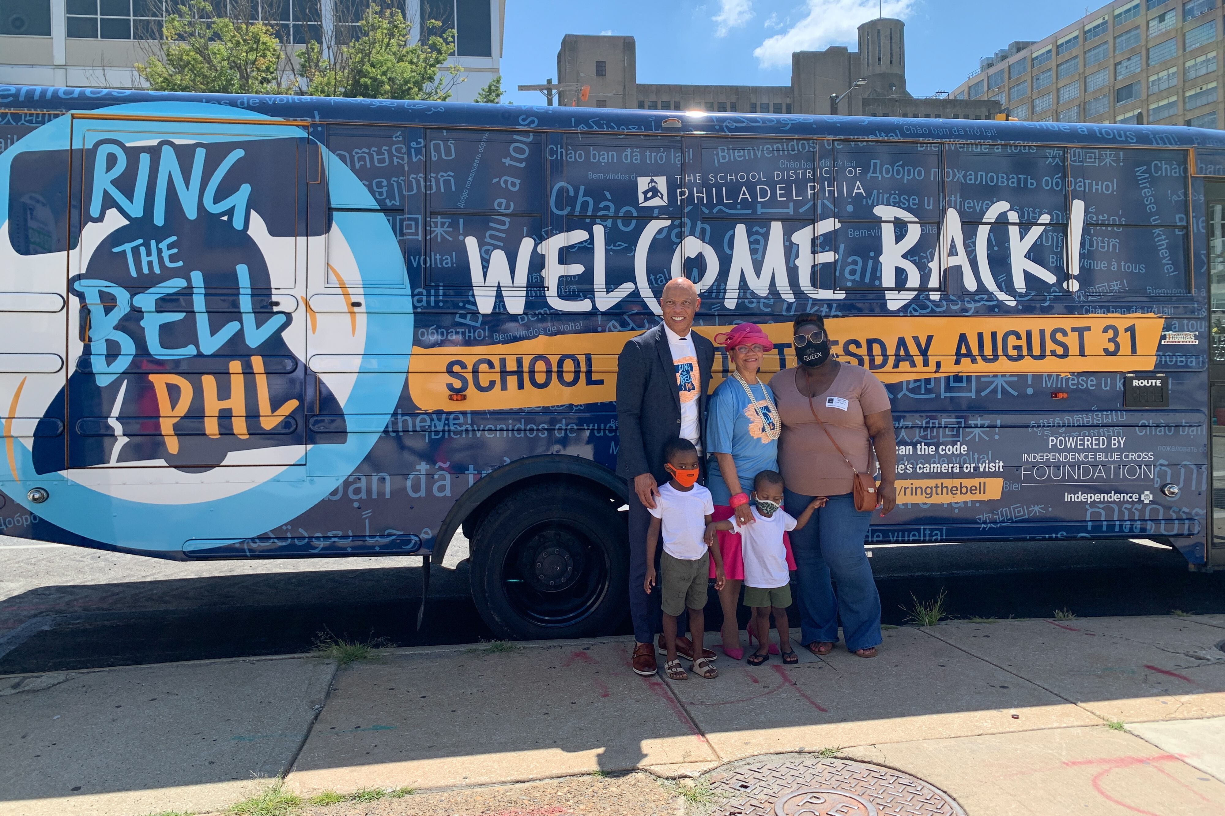 Philadelphia Superintendent William Hite stands in front of a large bus that reads “Ring the Bell PHL, Welcome Back!” with two women and two young boys.