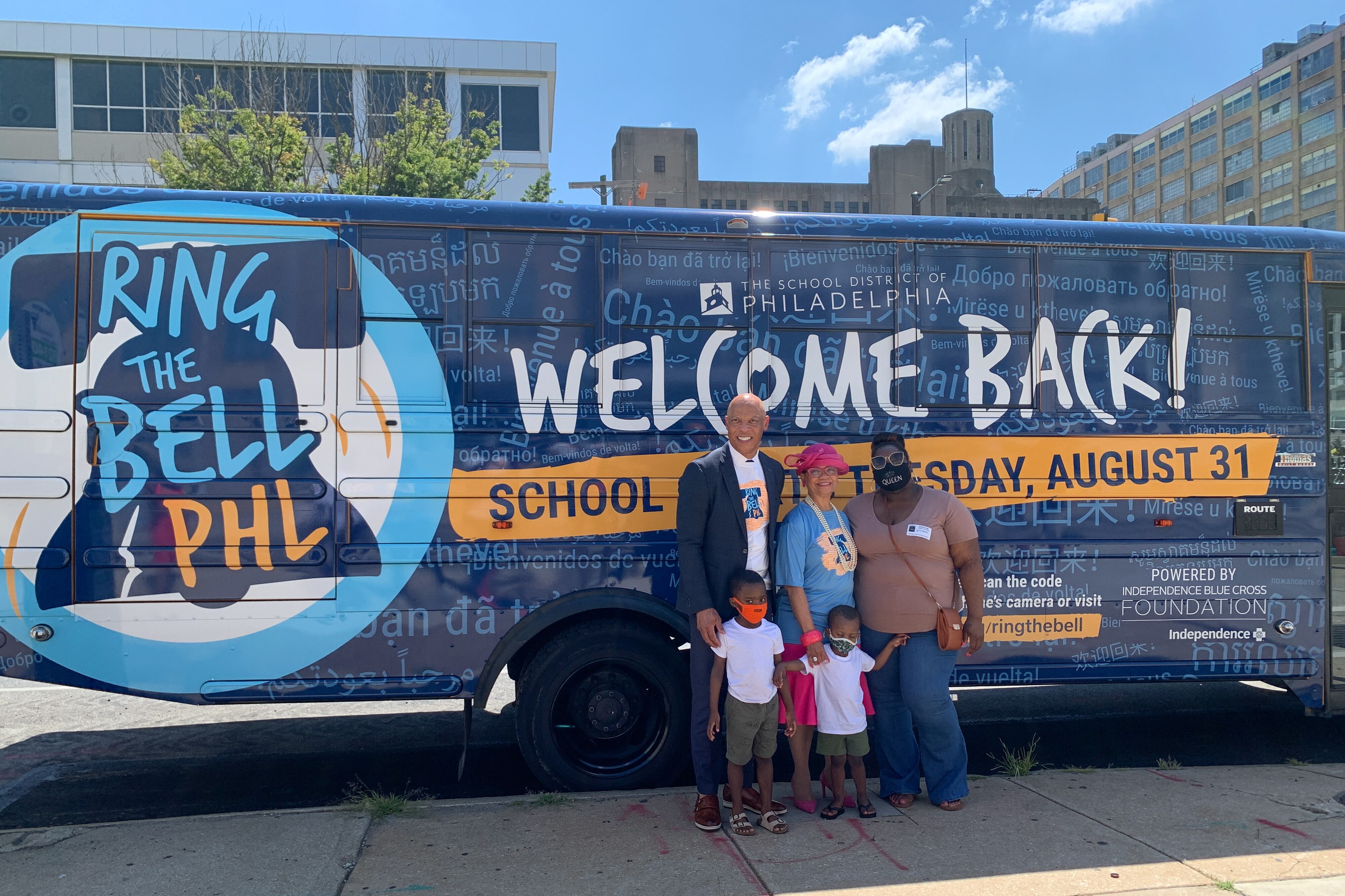 Philadelphia Superintendent William Hite stands in front of a large bus that reads “Ring the Bell PHL, Welcome Back!” with two women and two young boys.