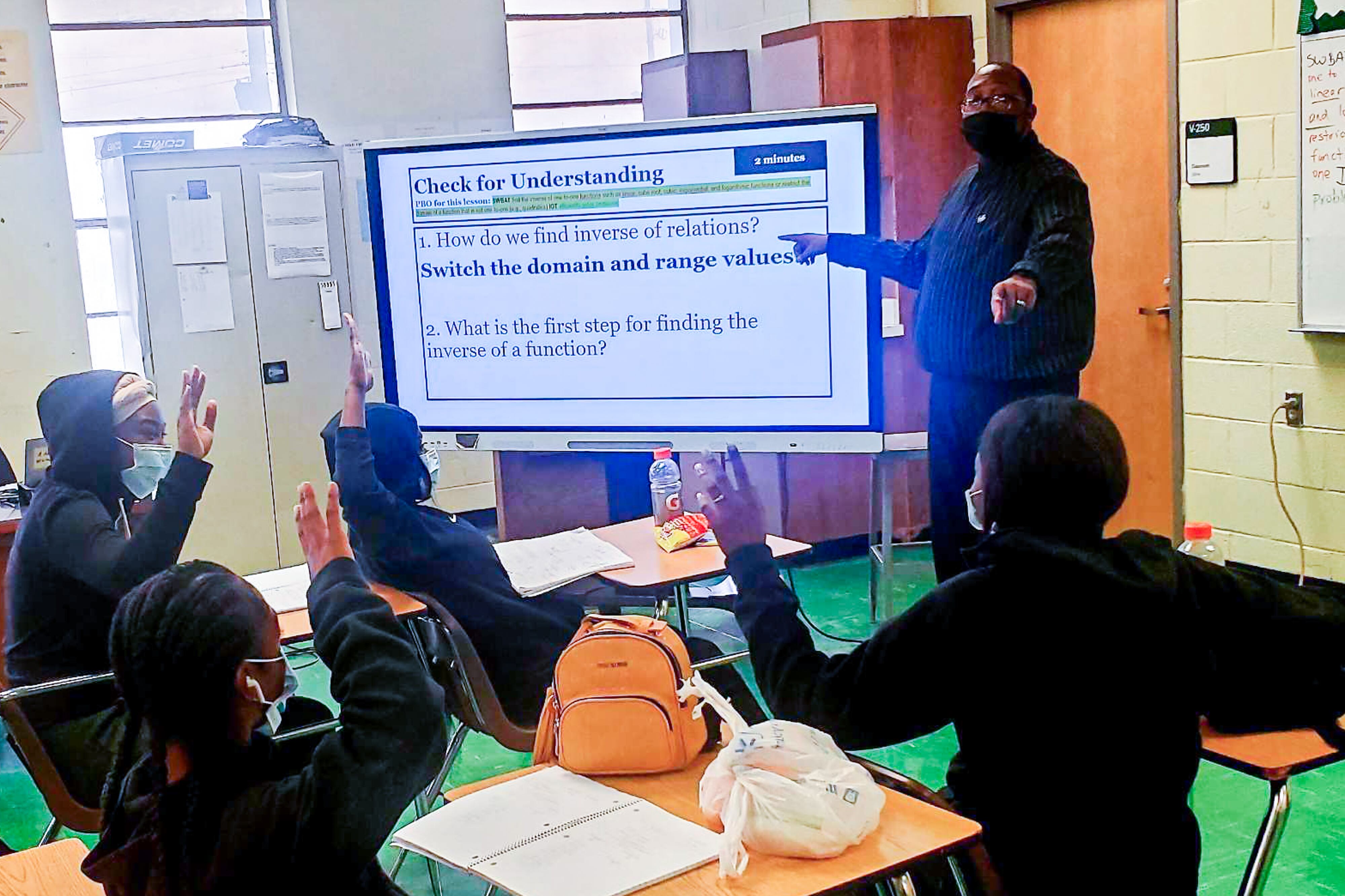 A male teacher stands in a classroom in front of a large monitor as engaged students watch from their desks and raise their hands to contribute to the discussion.
