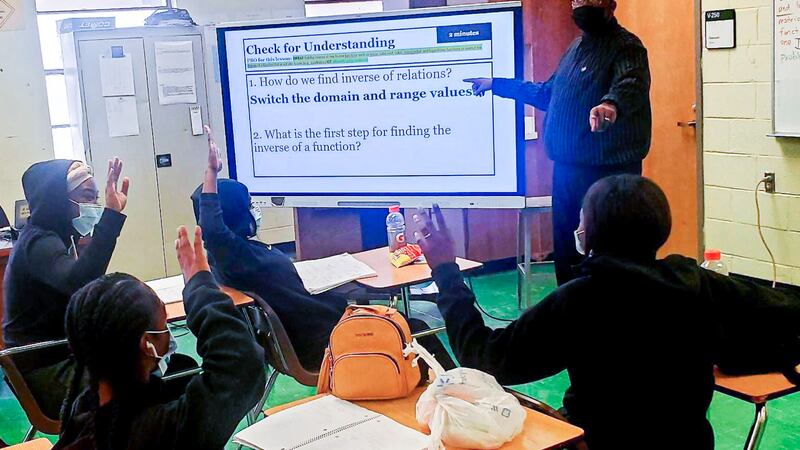A male teacher stands in a classroom in front of a large monitor as engaged students watch from their desks and raise their hands to contribute to the discussion. 