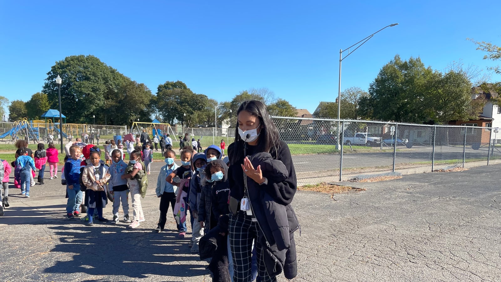 A young woman leads her kindergarteners, who are walking in a line behind her on a blacktop.