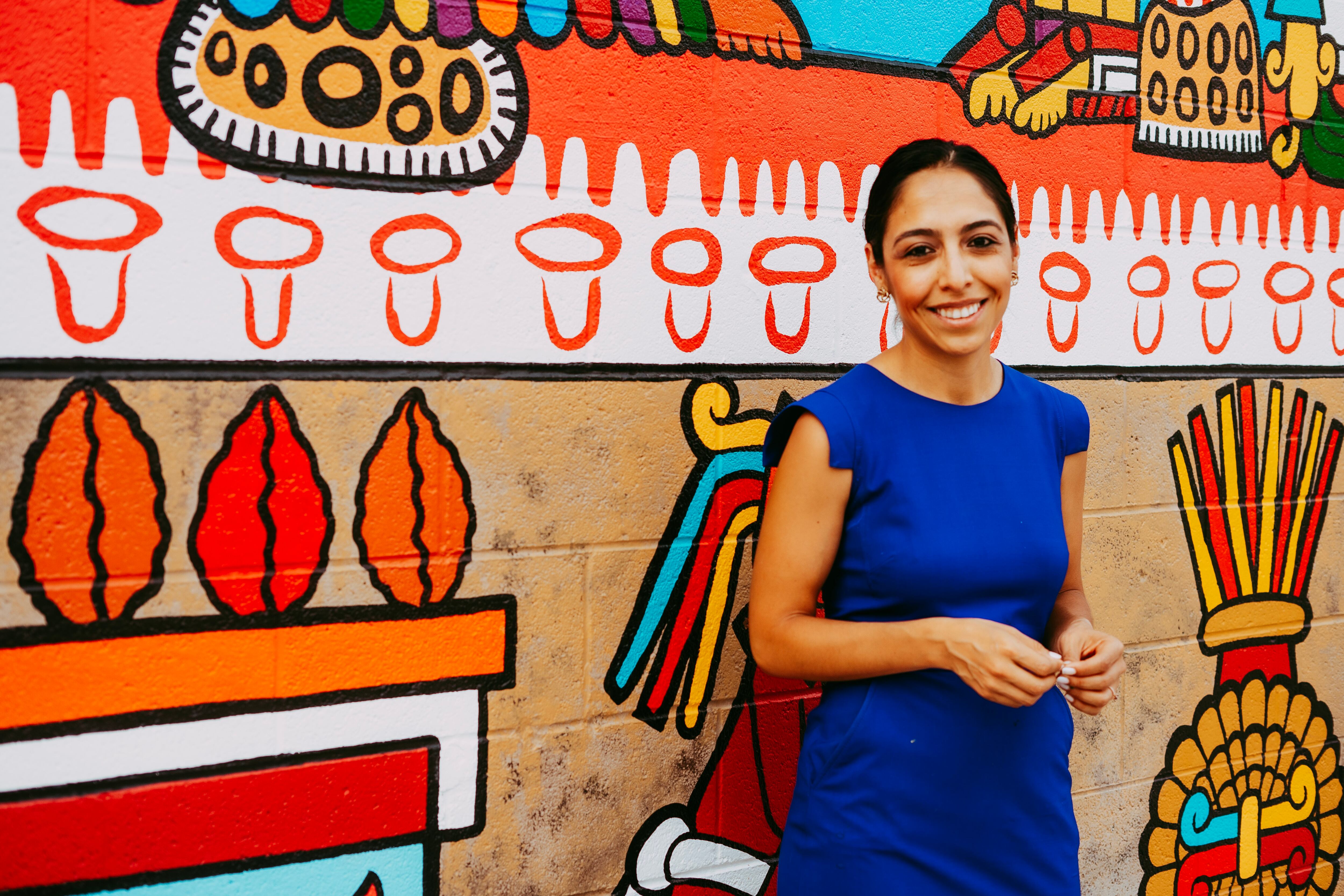A woman wearing a blue dress poses in front of a brightly colored mural.
