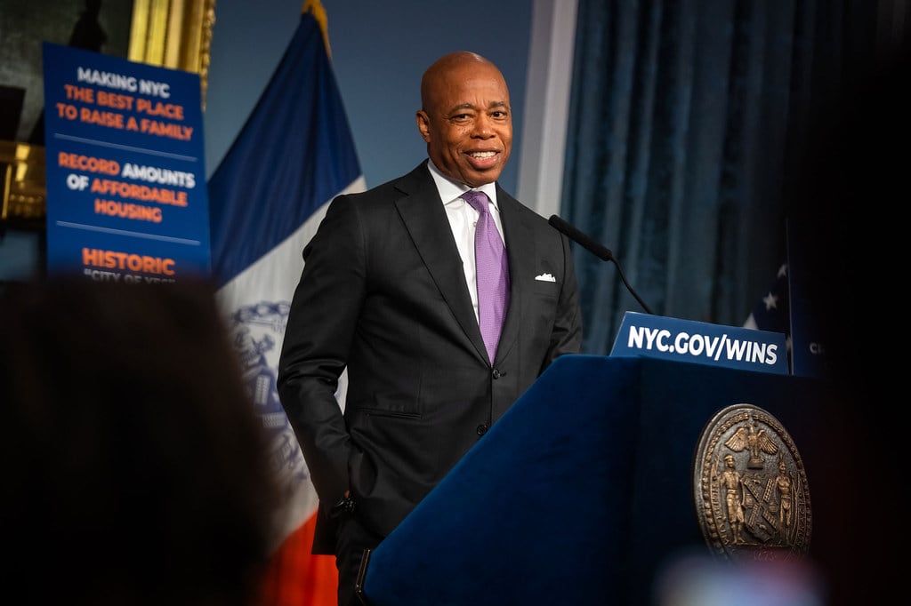 A man in a dark suit and lavender tie speaks behind a podium