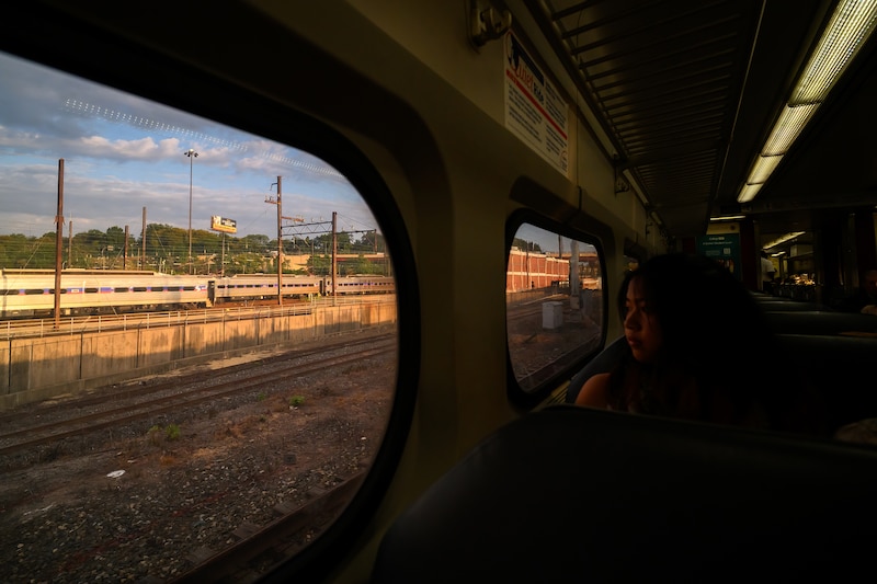 A photograph of a high school girl sitting in a darkly lit train car with a large outside scene in the window.