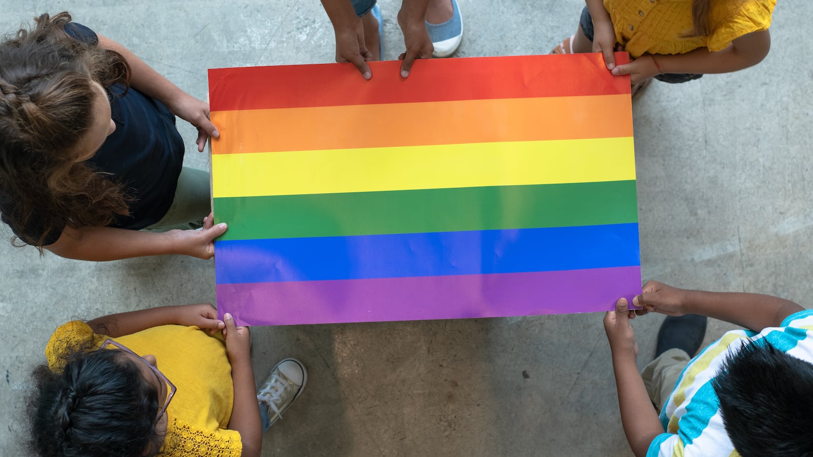 A top-down view of a rainbow-colored flag held at the edges by five people.