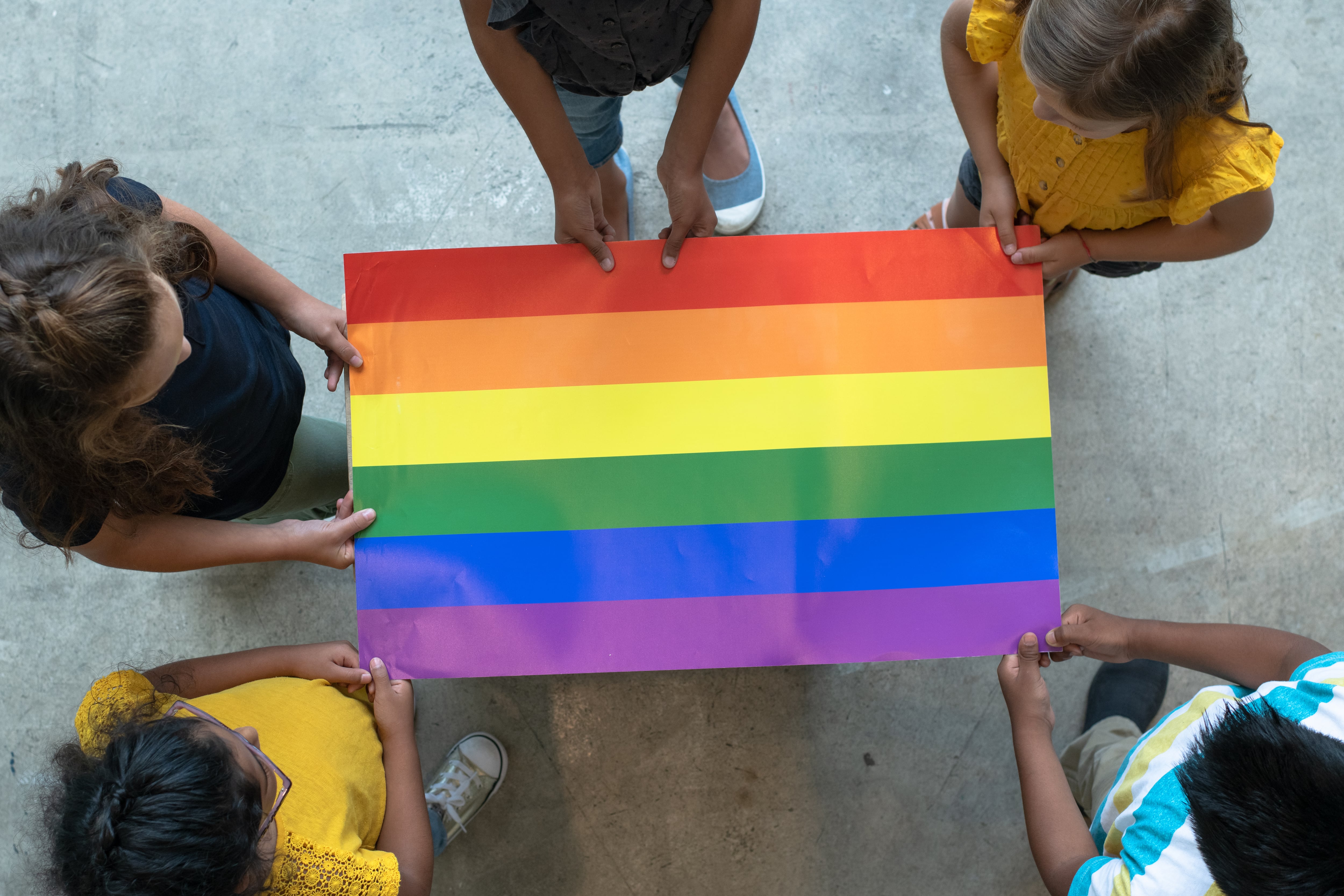 A top-down view of a rainbow-colored flag held at the edges by five people.