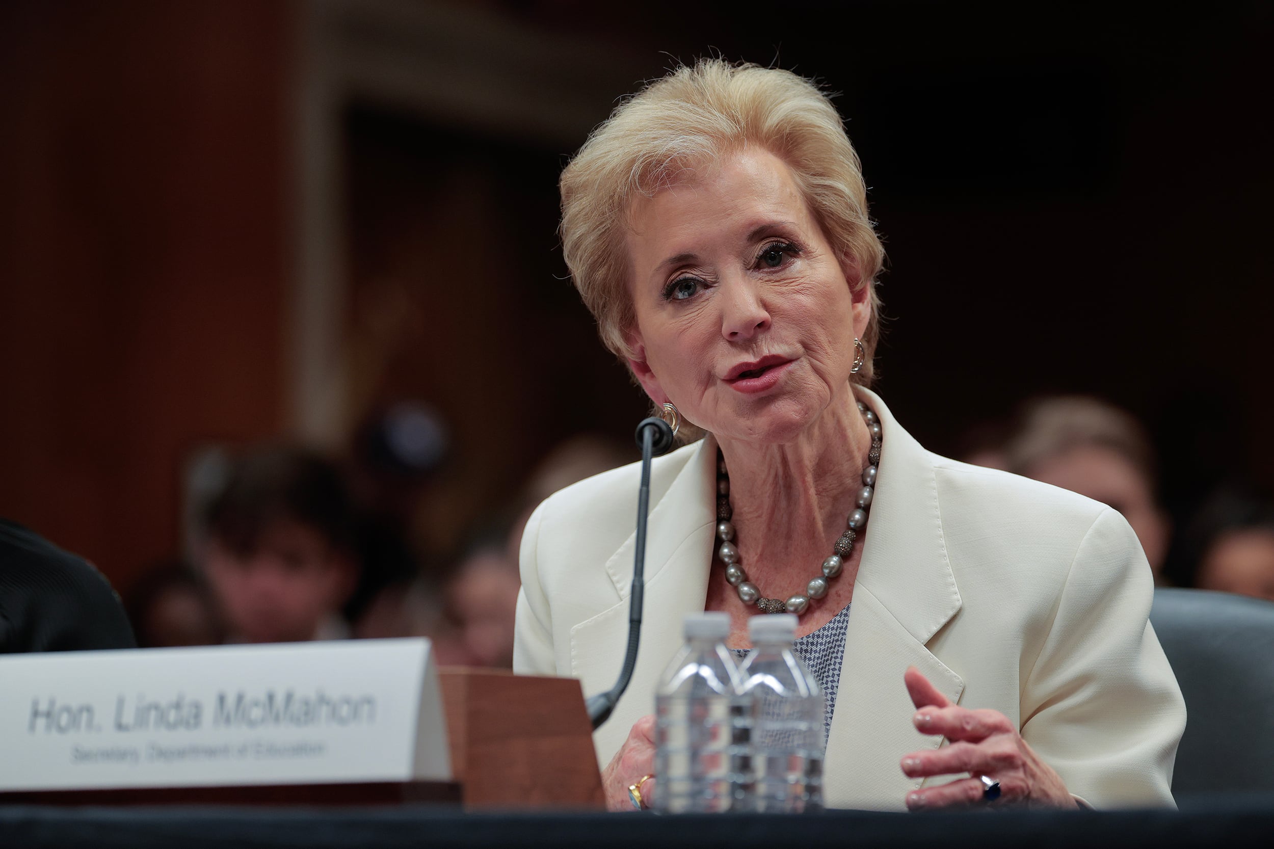 A photograph of a white woman wearing a white suit jacket speaks at a wooden desk.