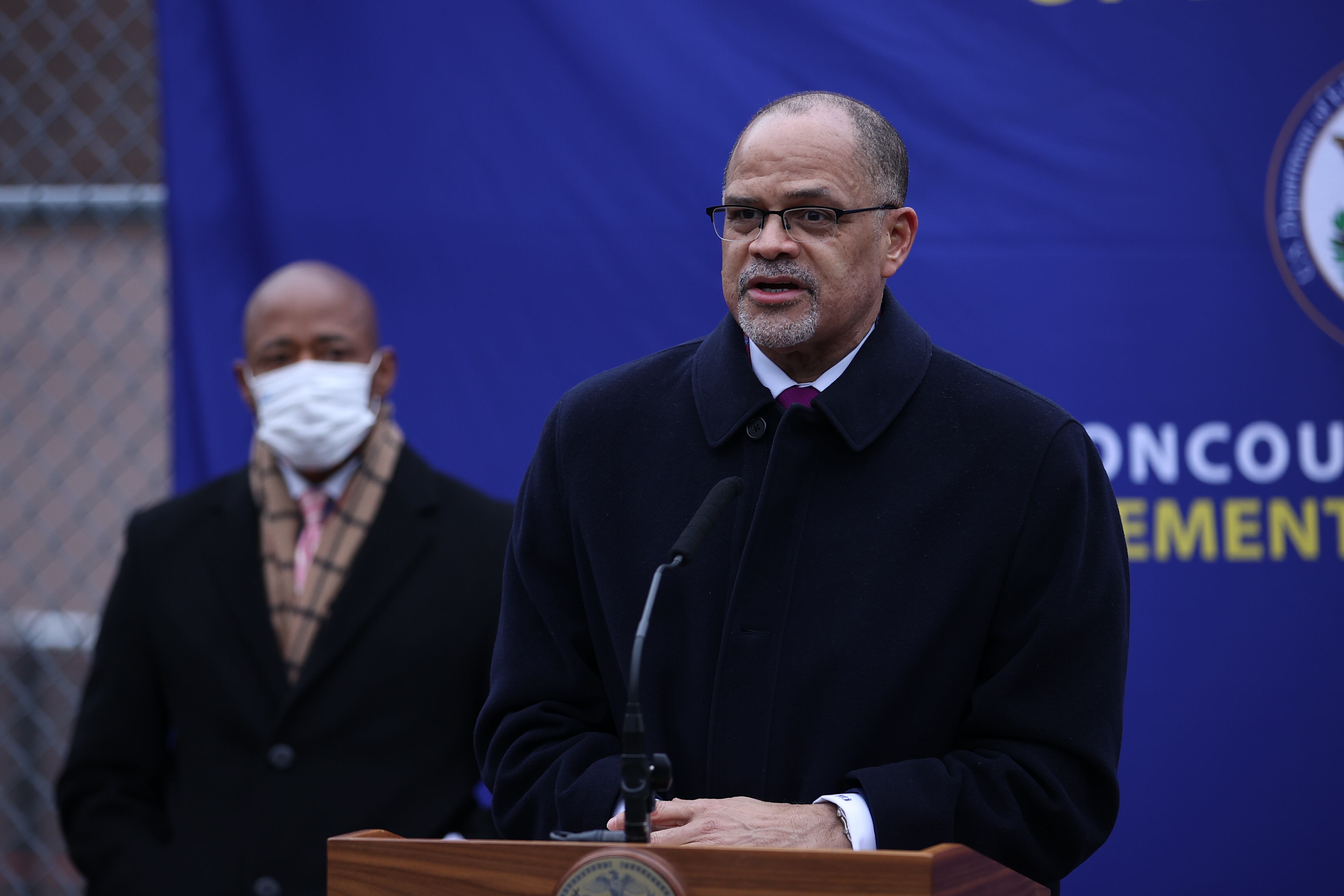 A man wearing a black coat stands outside at a lectern.