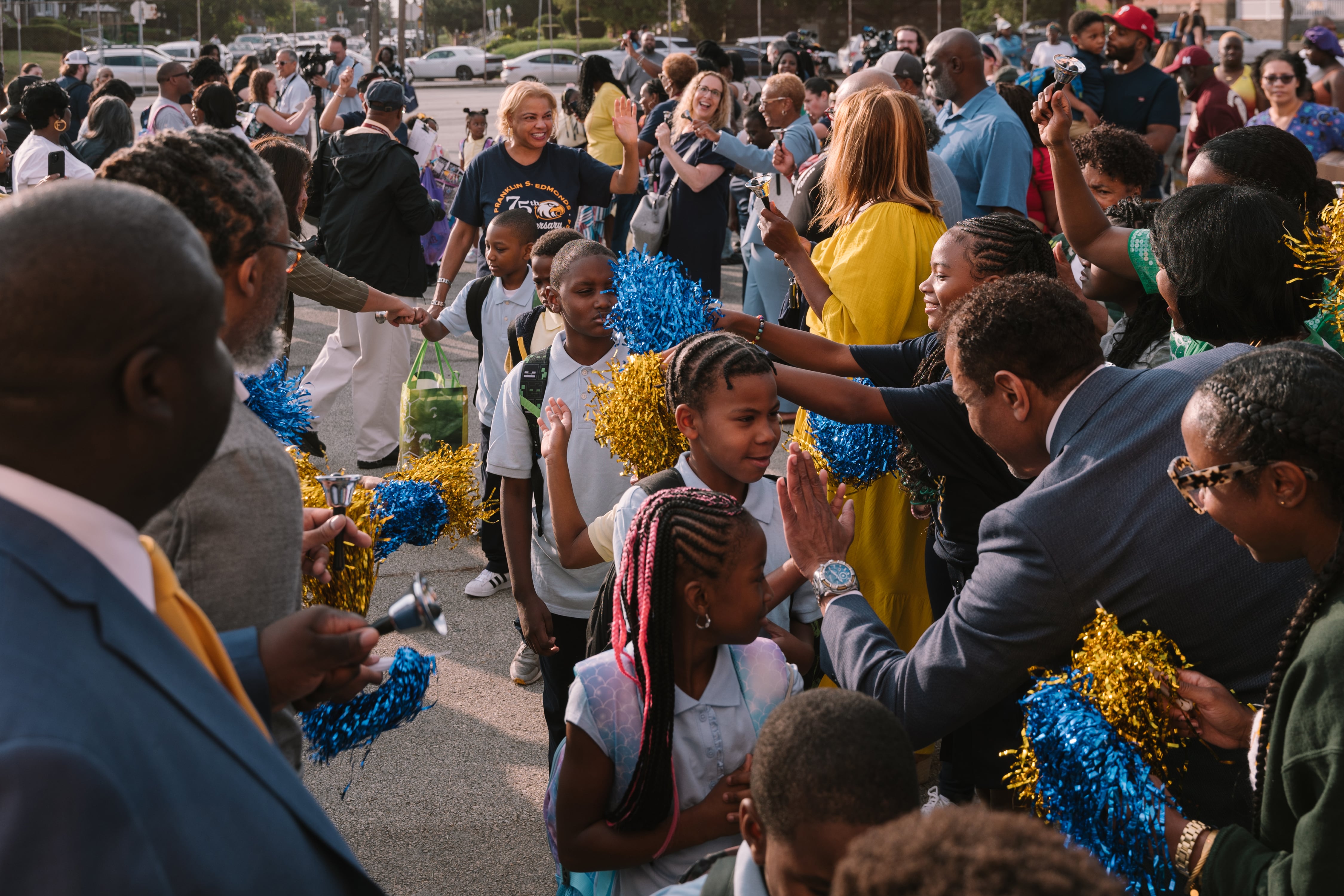A large group of young students walk in a line between two groups of adults high-fiving and waving on the first day of school.
