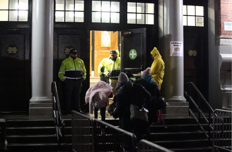 A group of people in rain jackets enter a school building as two guards are at the door.