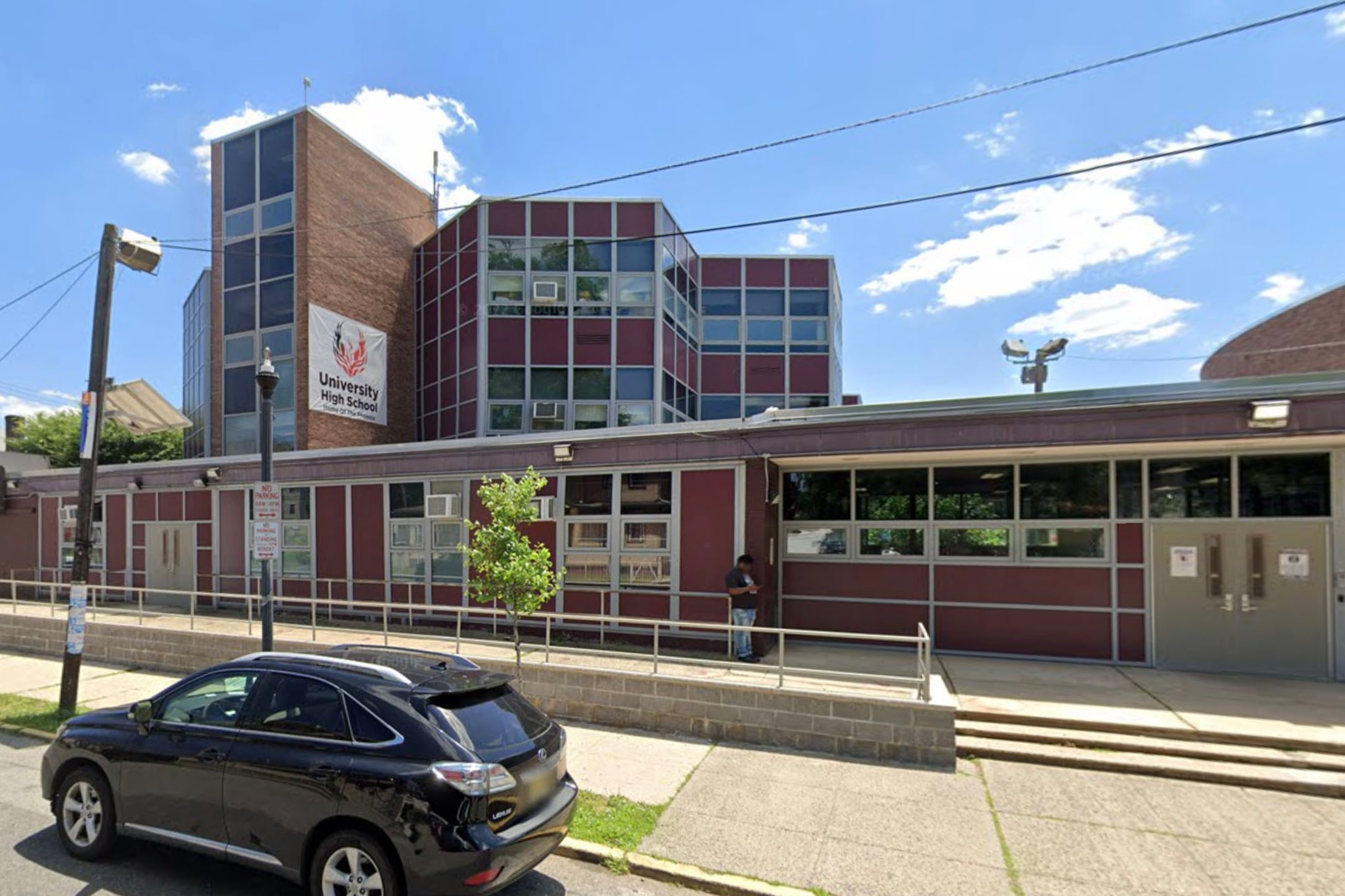 A large red brick school building with a car parked on the street in front and blue sky with clouds in the background.