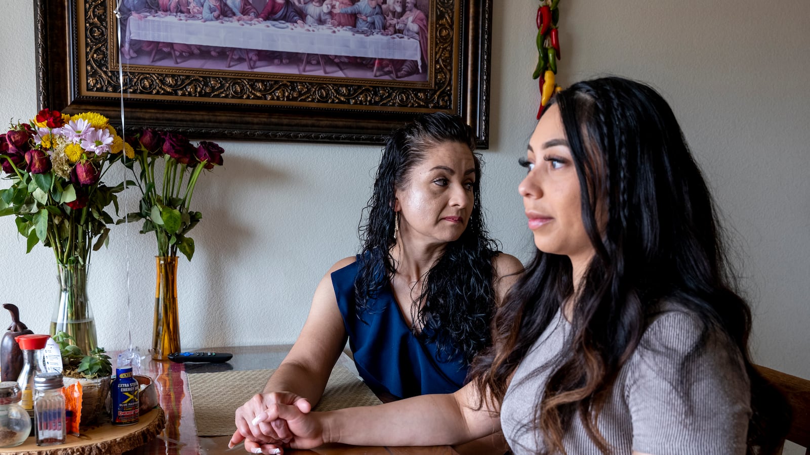 A mother holds her adult daughter’s hand as they sit at the family’s kitchen table.