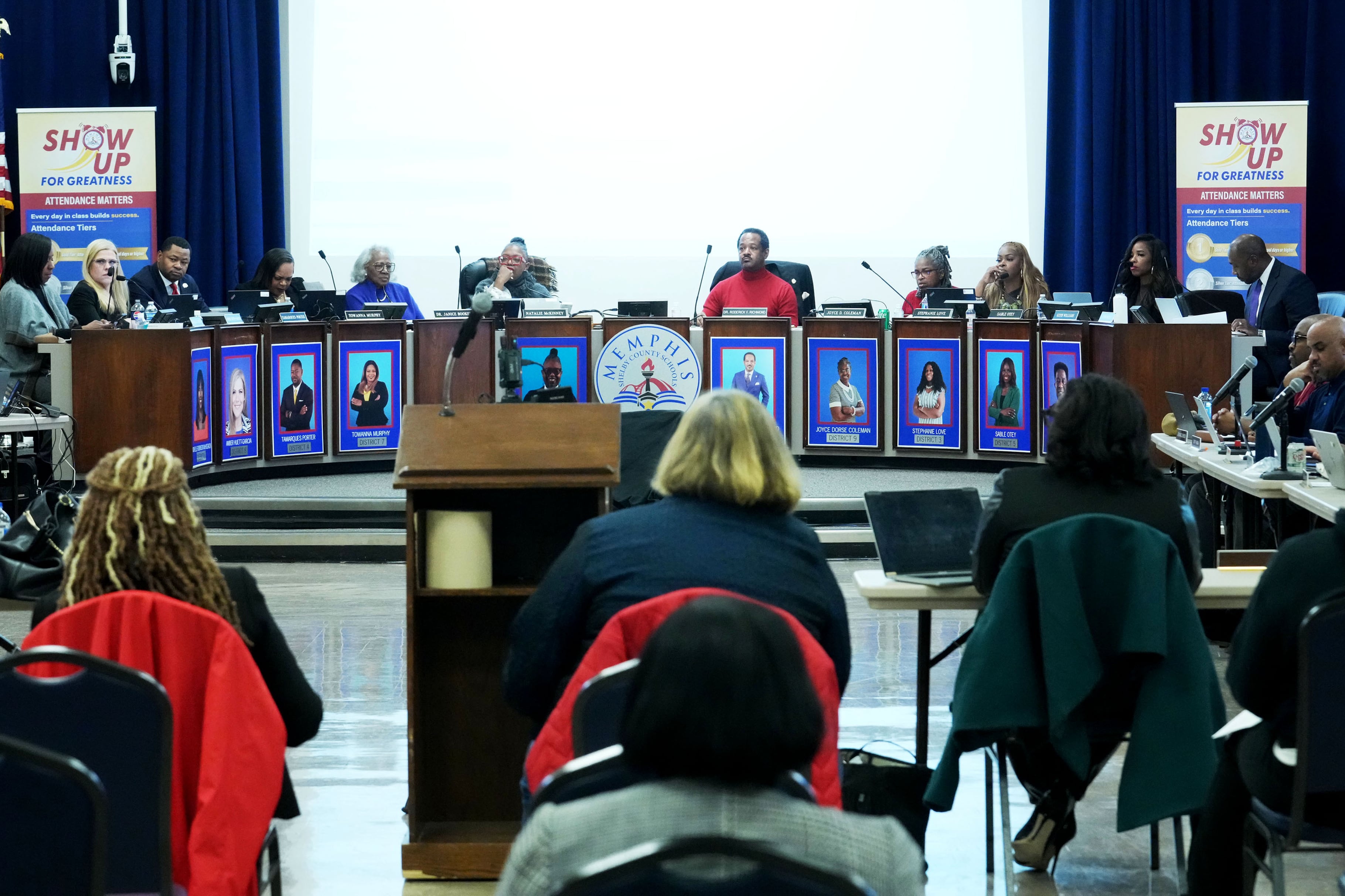 A photograph of a large conference room with a group of adults sitting at a large table in the background in focus while a large group of community members sit in chairs facing them.