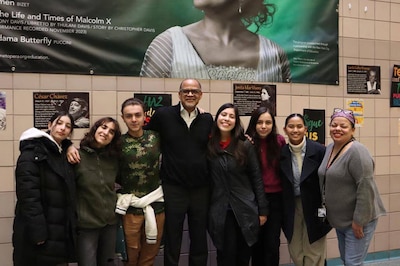 Eight people stand in a line posing for a photograph. There is a large poster hanging on a tan tile wall in the background.