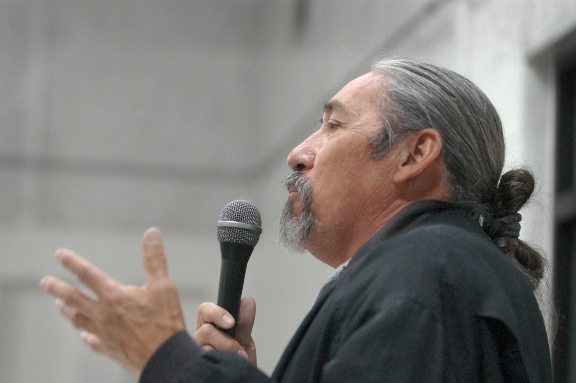 A middle-aged man dressed in black, with long gray hair pulled back into a knot, speaks into a microphone. He’s seen in profile, facing left, and he’s gesturing with his hand as he speaks.