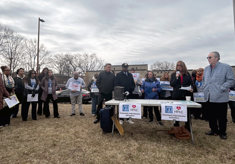 A group of people wearing coats stand outside, some holding signs and others speaking to a crowd.