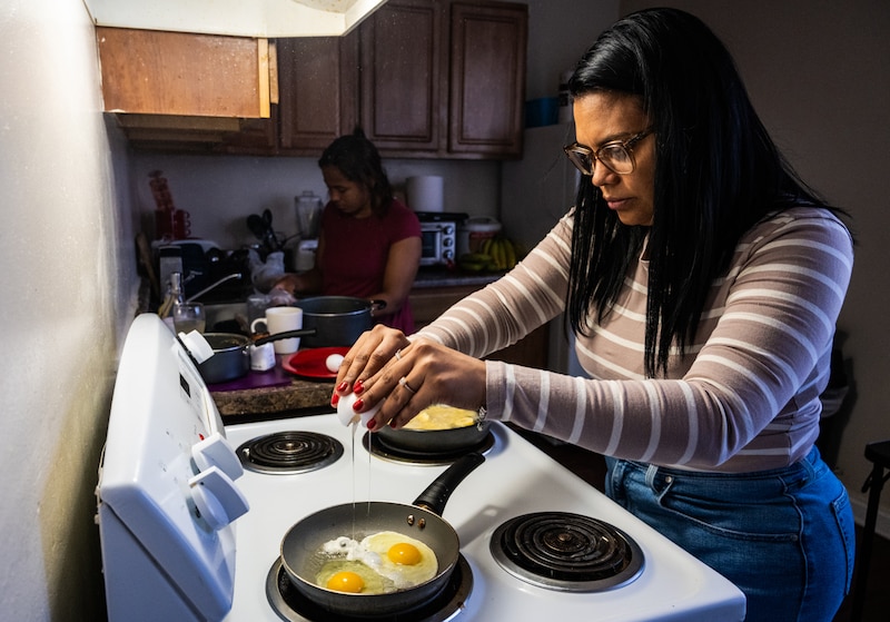An adult woman with long dark hair cracks an egg over a frying pan at an over with another person out of focus in the background of the kitchen.