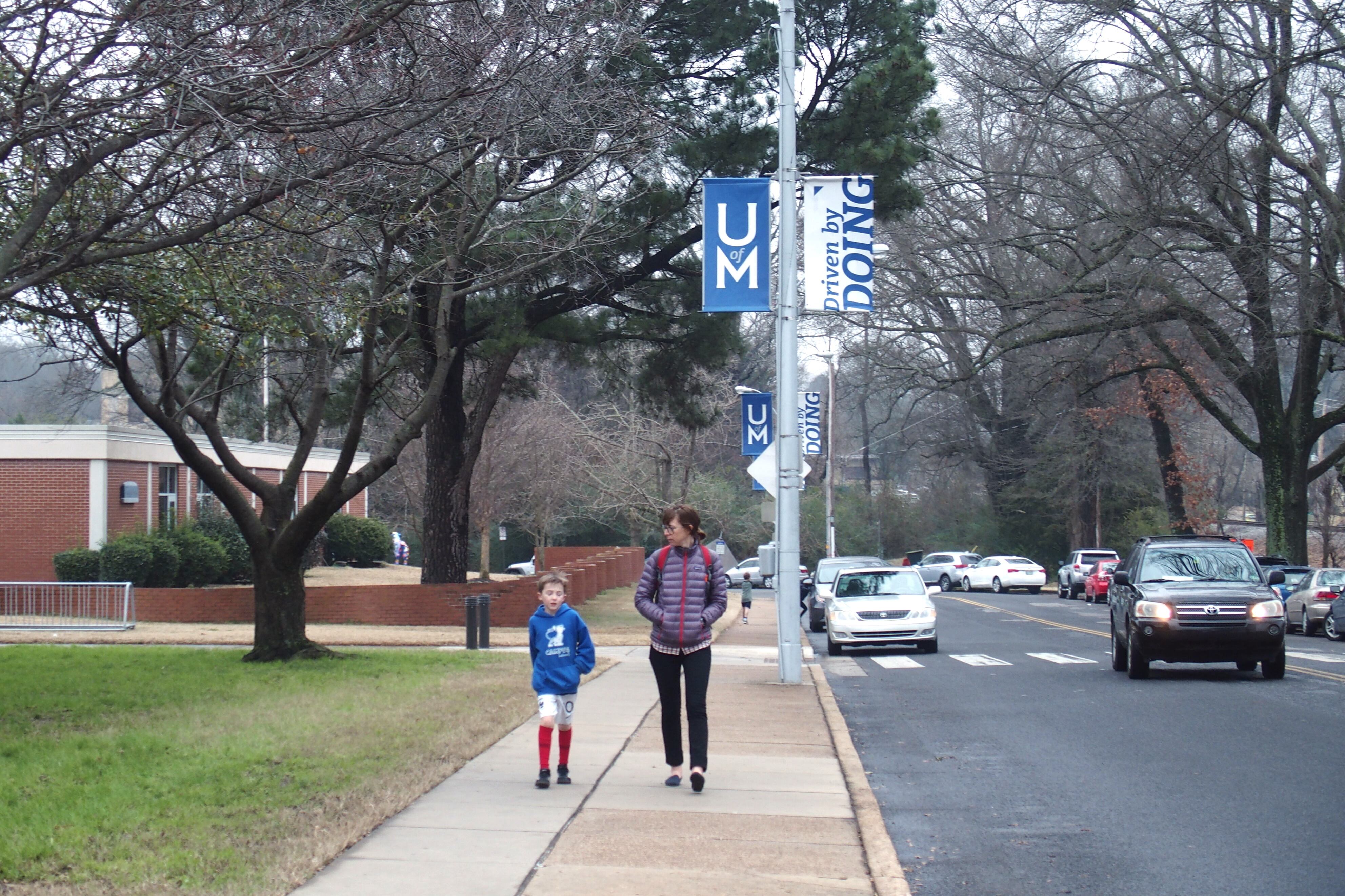 A young student walks with another person outside a school next to a road with cars driving along.