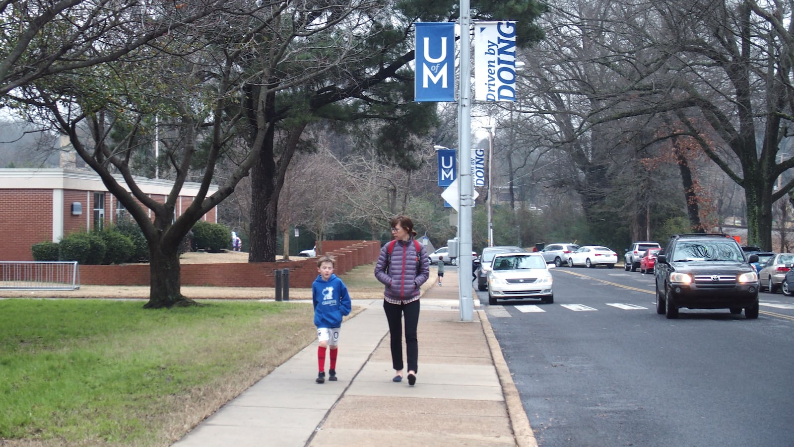 A young student walks with another person outside a school next to a road with cars driving along.
