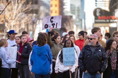 A photograph of a large group of students marching on a sunny day, some are holding protest signs.
