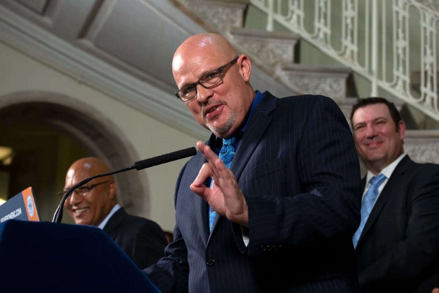 A man with glasses in a black blazer and blue shirt stands at a podium.