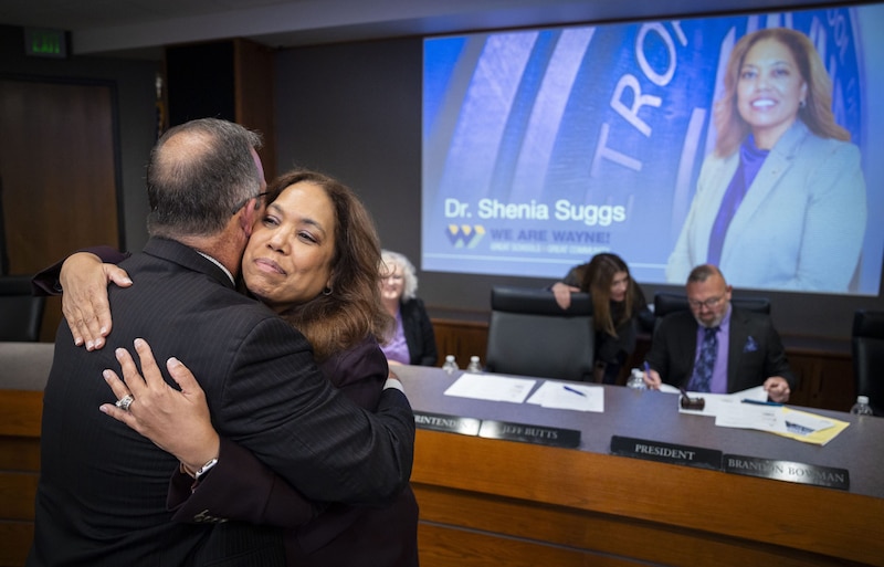 A photograph of a man and woman in suits hugging in front of a large screen.